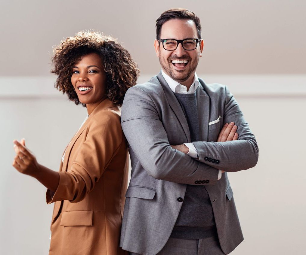 A professional man and woman standing back-to-back, smiling, against a neutral background.