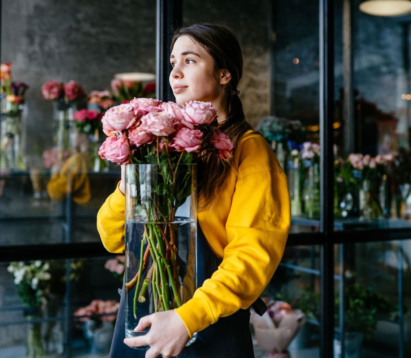A person wearing a yellow sweater holds a tall glass vase of pink roses in a flower shop.