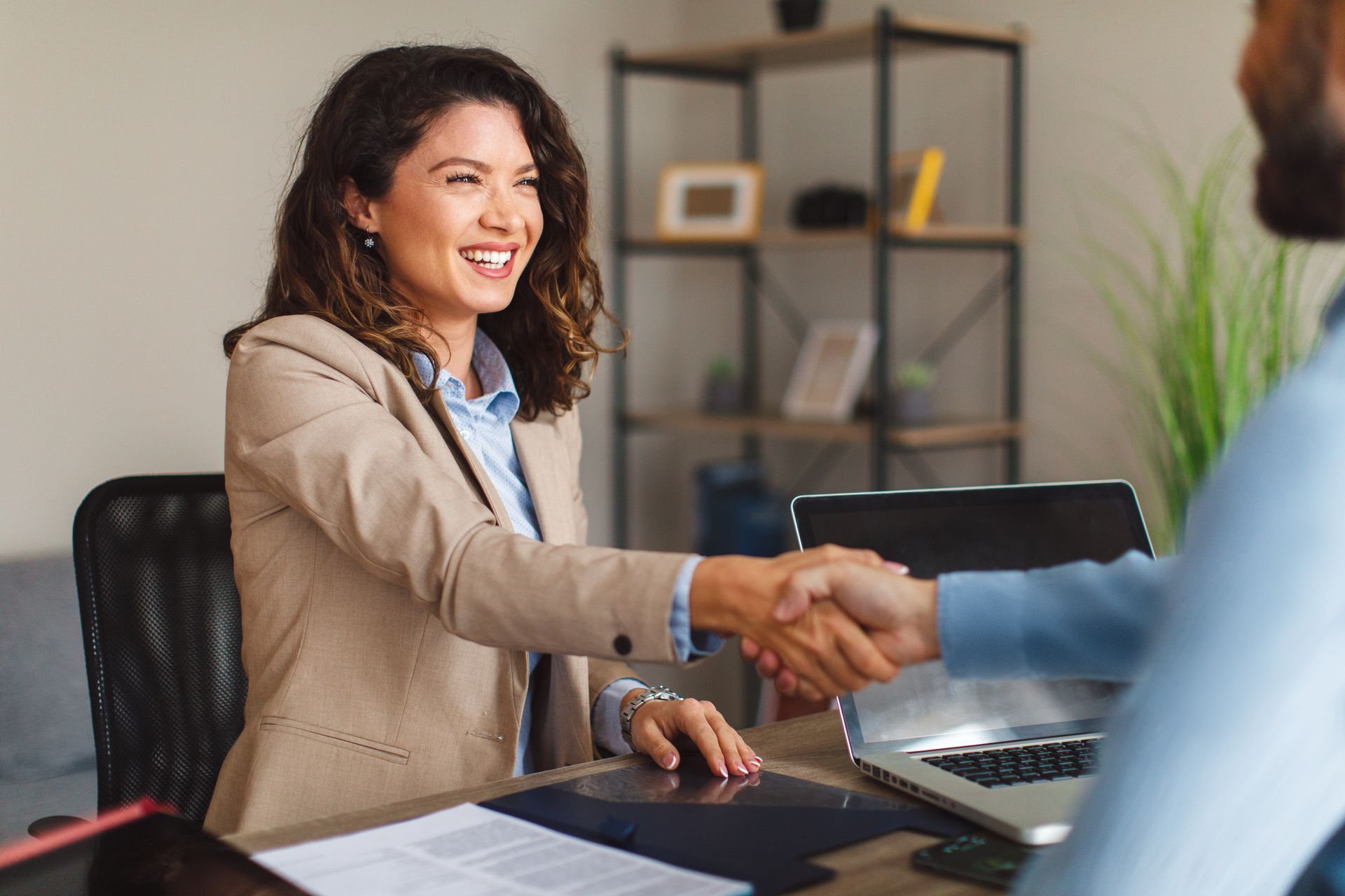 A smiling professional shakes hands with someone across a desk in a brightly lit office.