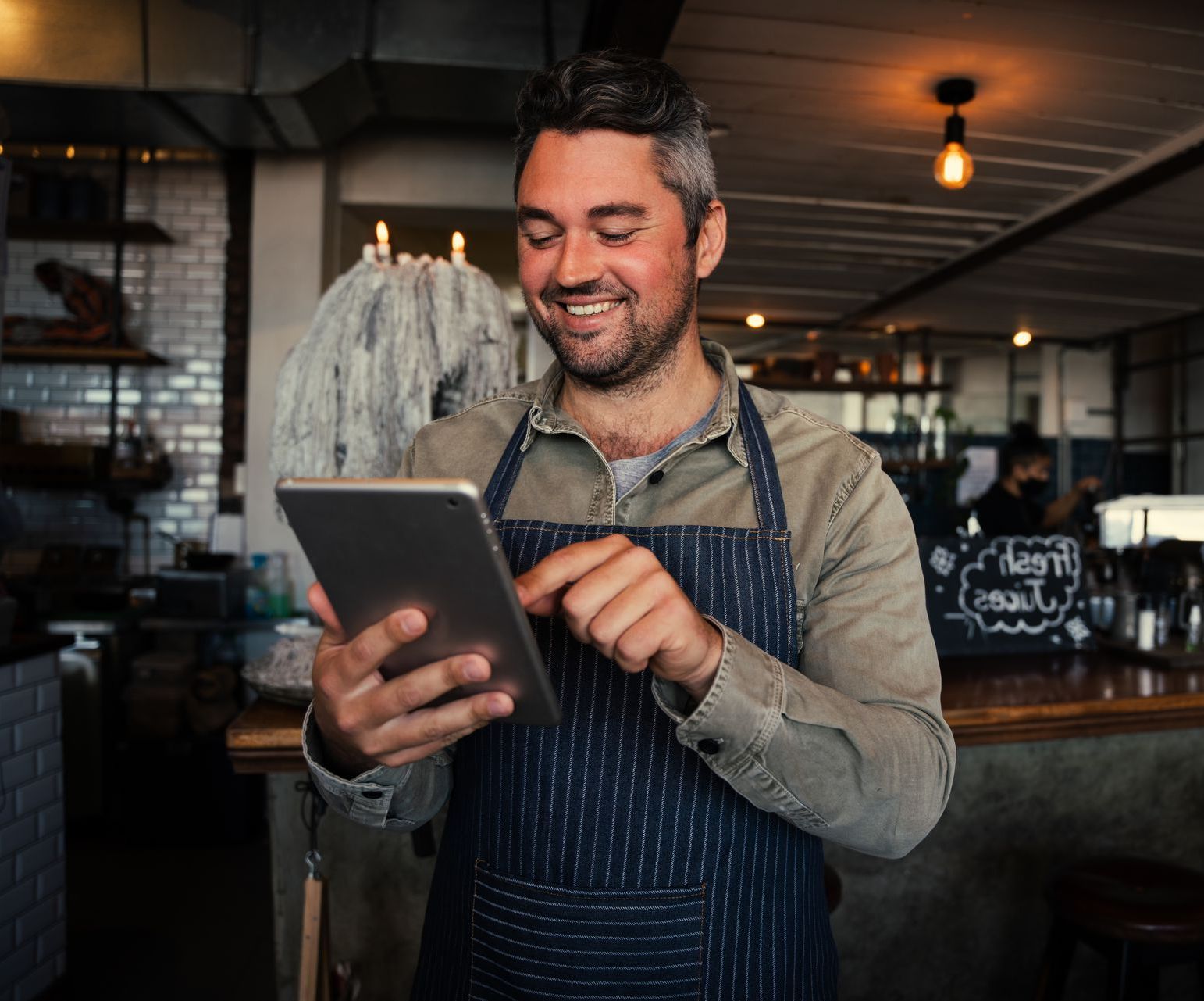 A smiling person wearing a dark denim apron uses a tablet while standing inside a restaurant or cafe.