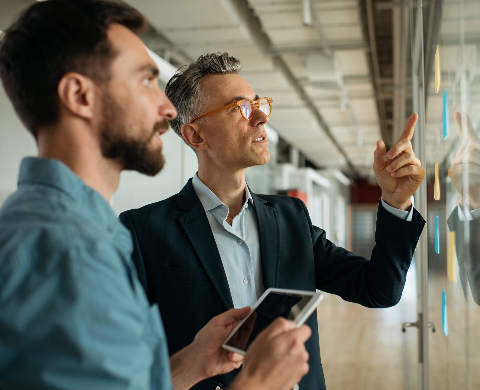 Two colleagues stand in an office, one holding a tablet and gesturing toward notes on a glass wall.