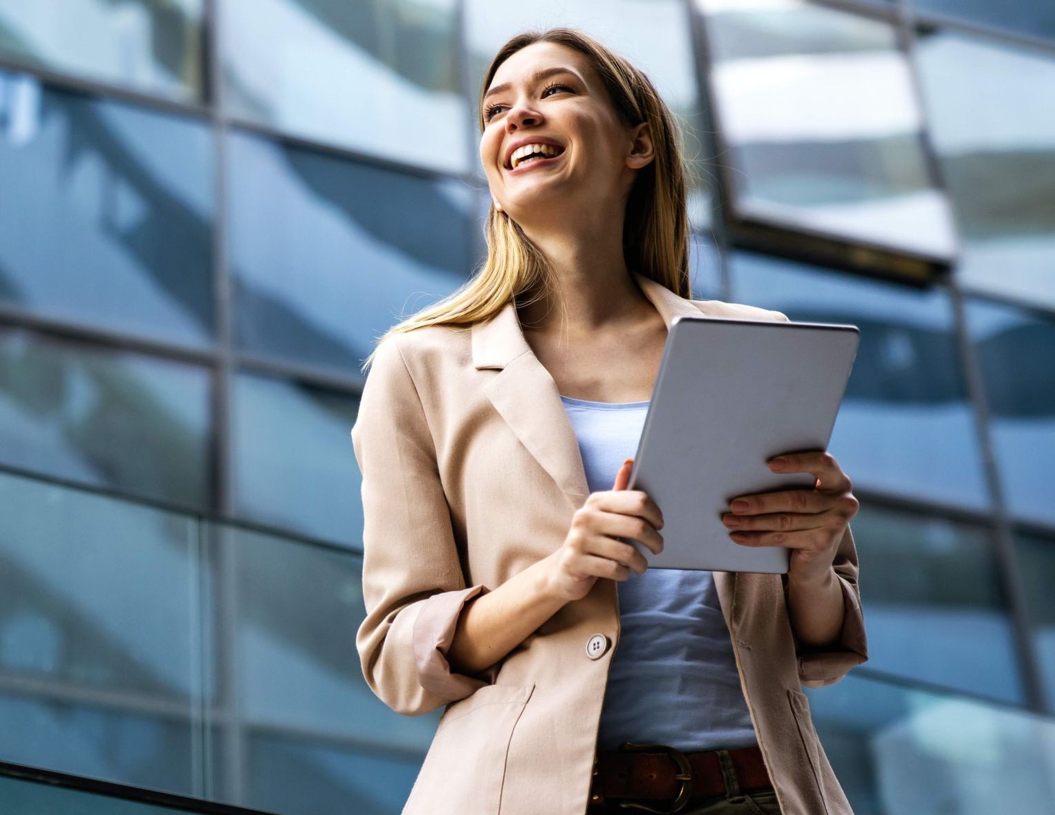 A smiling person in a beige blazer stands outdoors in front of a modern glass office building, holding a tablet.