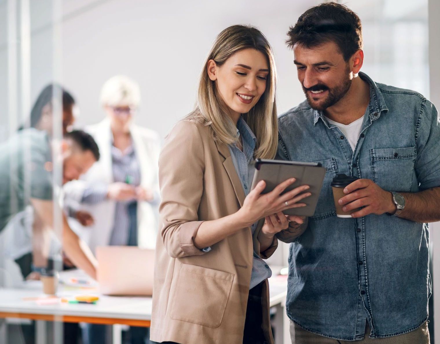 A smiling man and woman look at a tablet together in a bright, modern office with colleagues working in the background.
