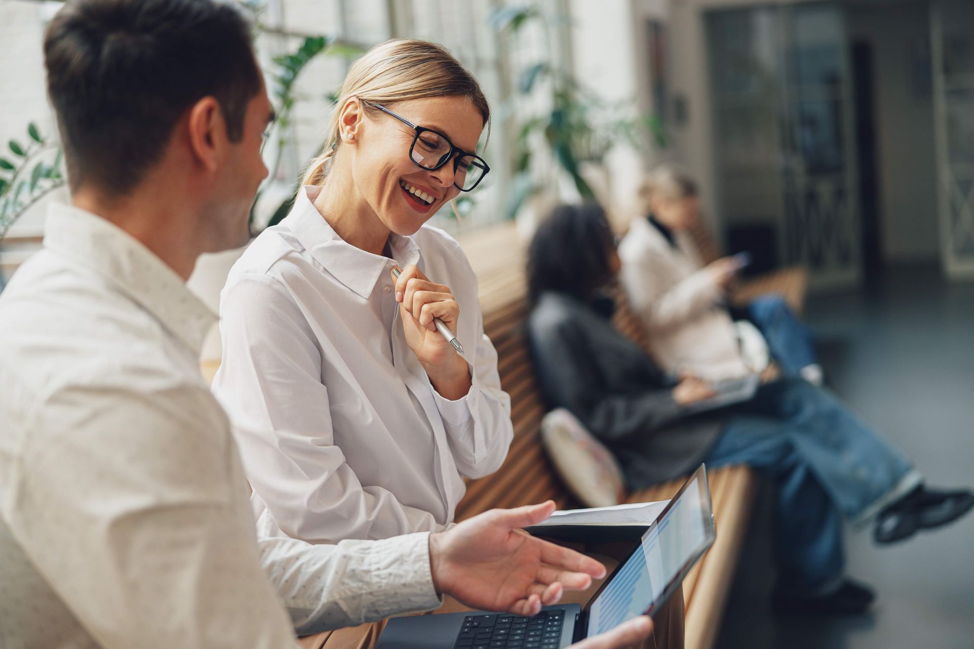 Two colleagues in white shirts sit on a wooden bench, smiling while reviewing information on an open laptop.