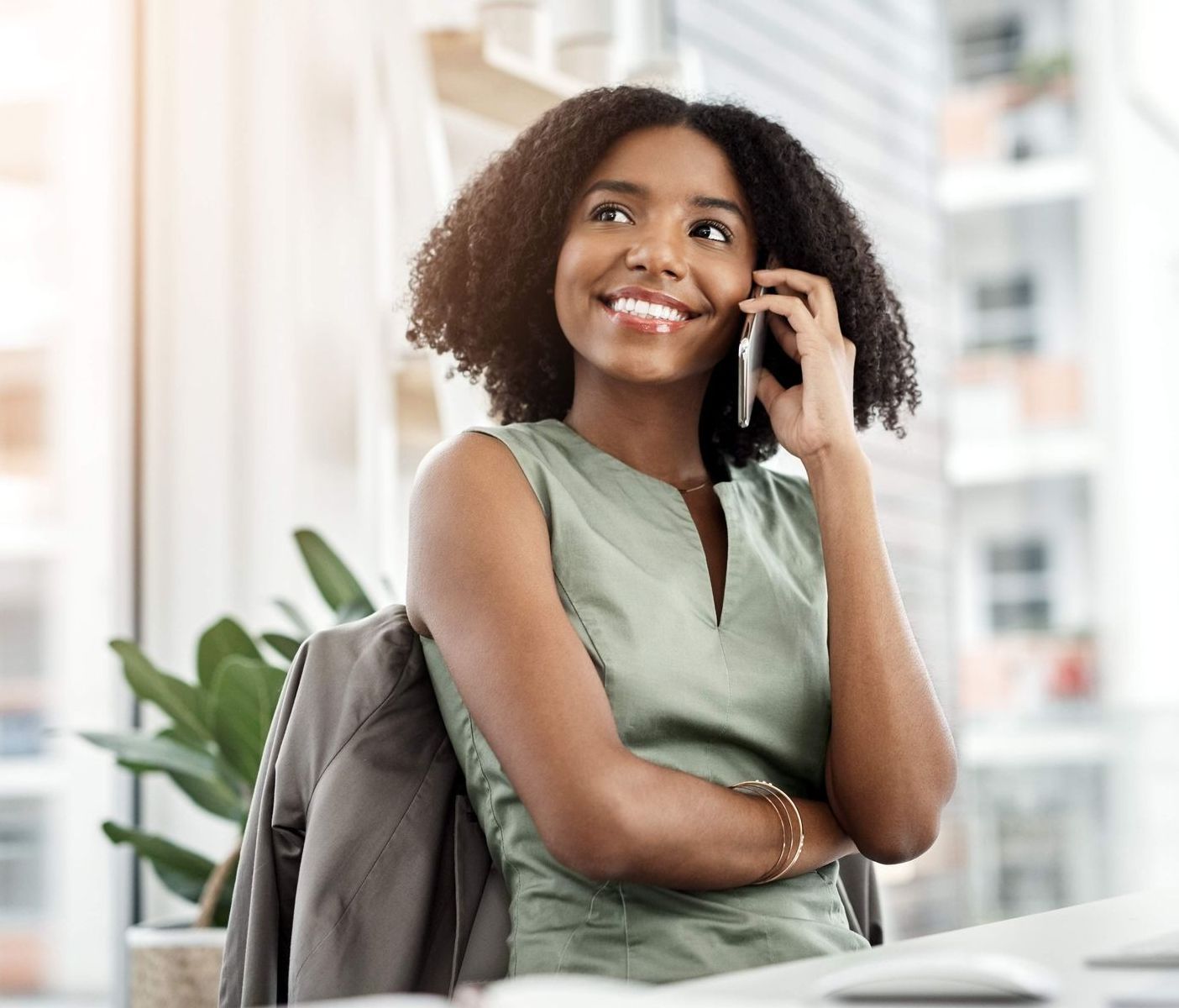 A smiling professional in a green top talks on a phone in a bright office, with a jacket draped over their chair.
