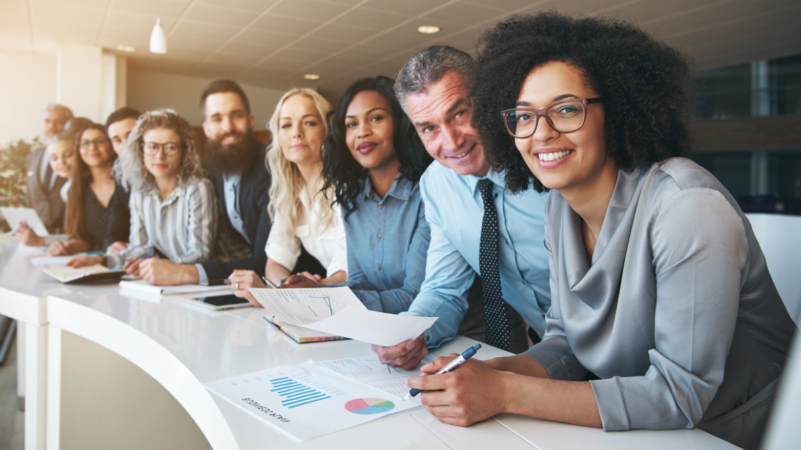 A diverse team sits at a curved desk in an office, smiling and reviewing documents and charts together.