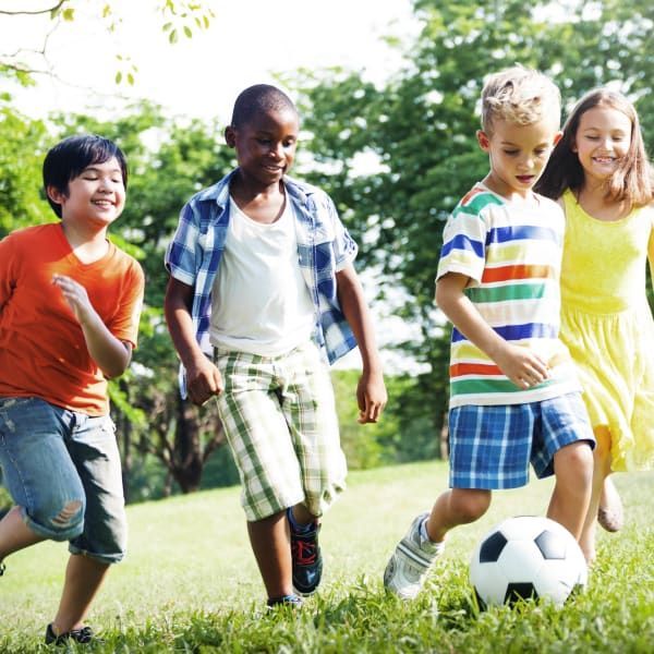 A group of four children play with a soccer ball in a grassy park, all smiling and running together outdoors.
