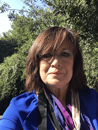 A woman in a blue shirt and purple scarf is standing in front of trees.