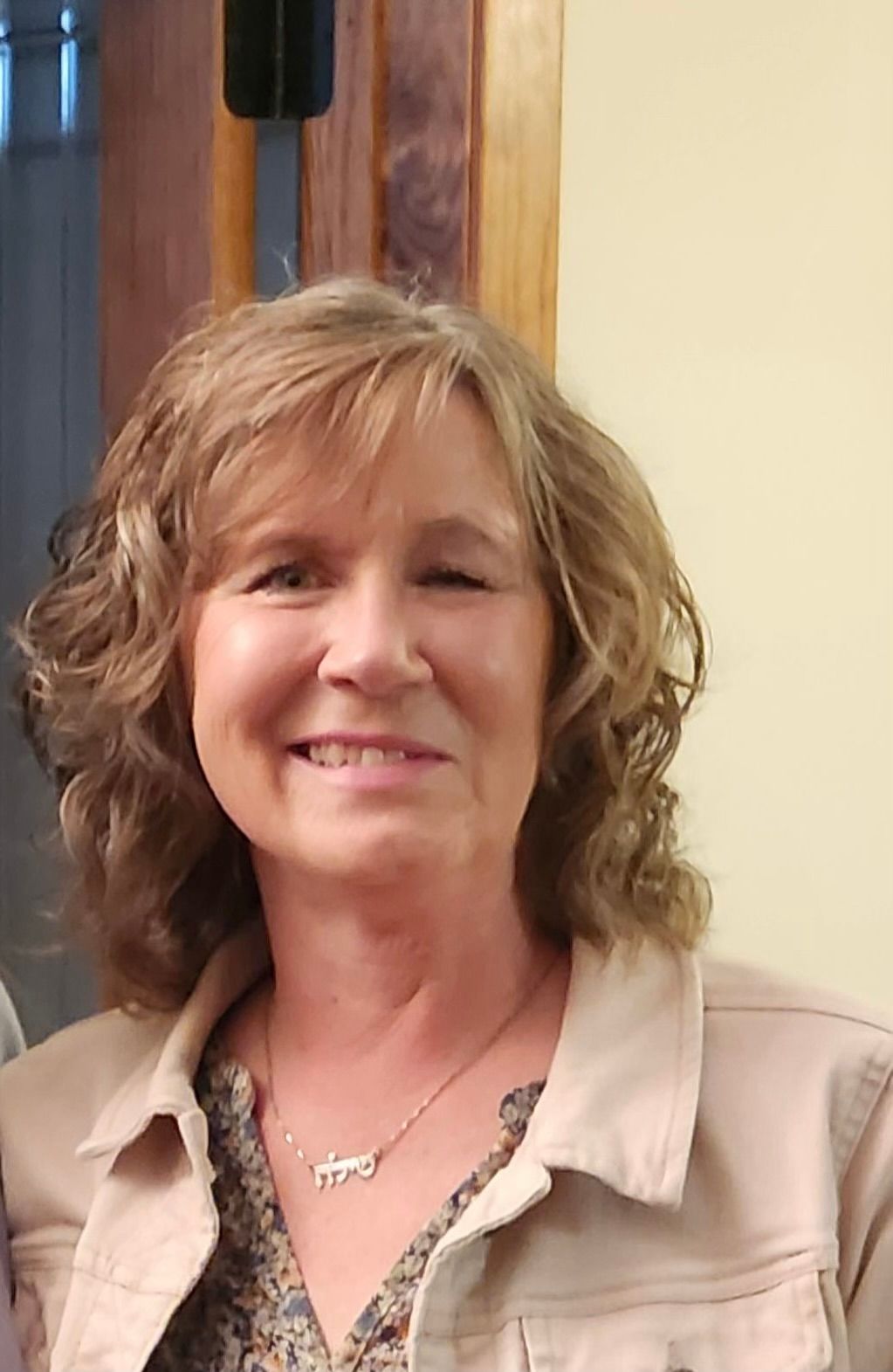 A woman wearing a tan jacket and a necklace is smiling for the camera.