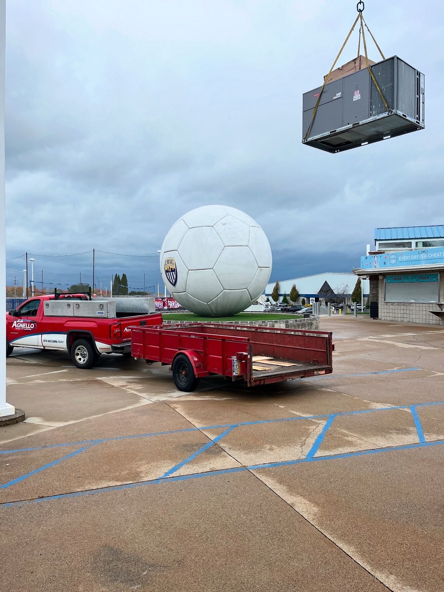 A red pickup truck tows a trailer behind a large soccer ball sculpture, while a crane lifts a rooftop HVAC unit nearby.