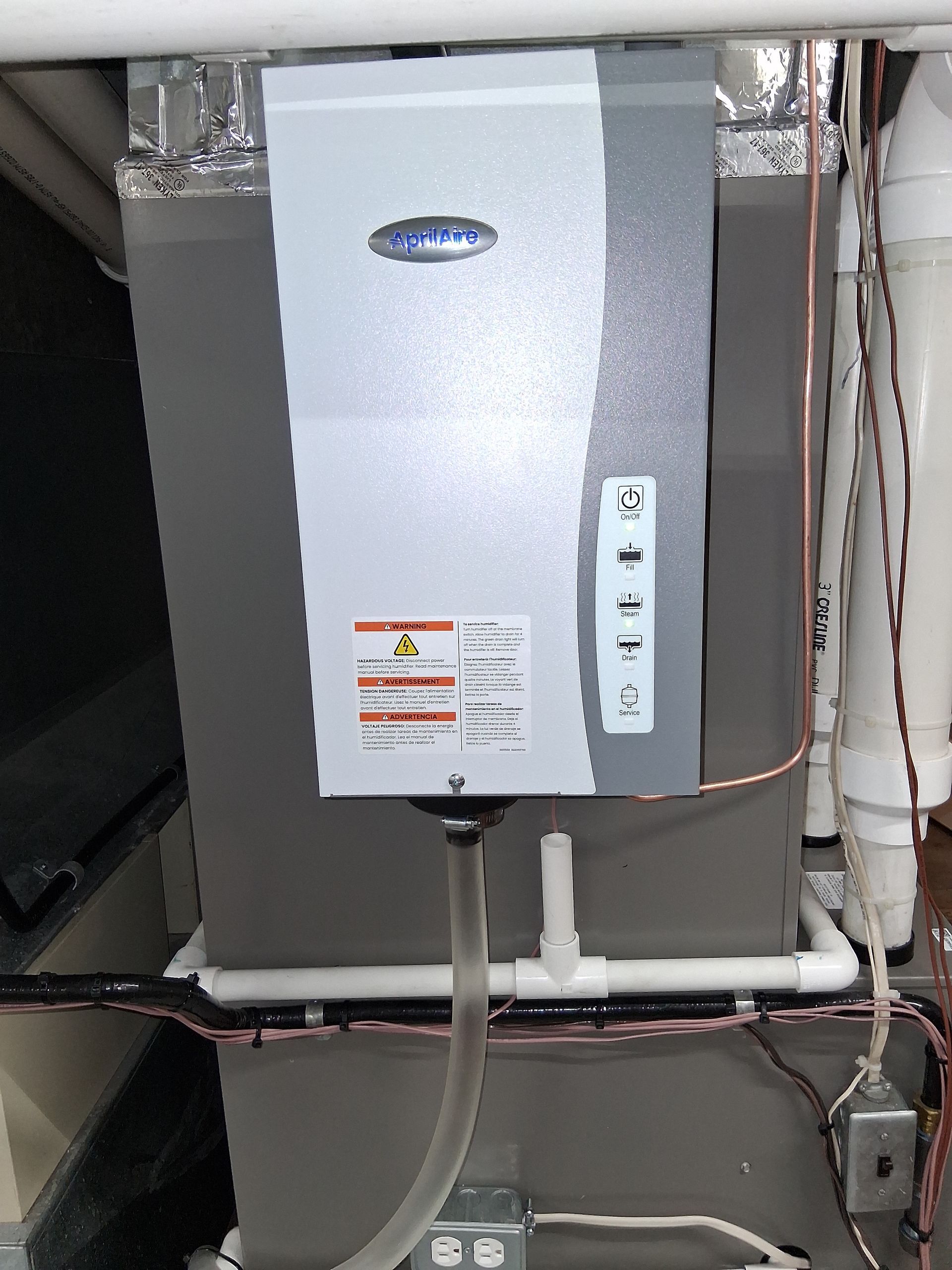 A gray Aprilaire humidifier mounted on the front of a gray HVAC furnace unit with connected piping and wiring.