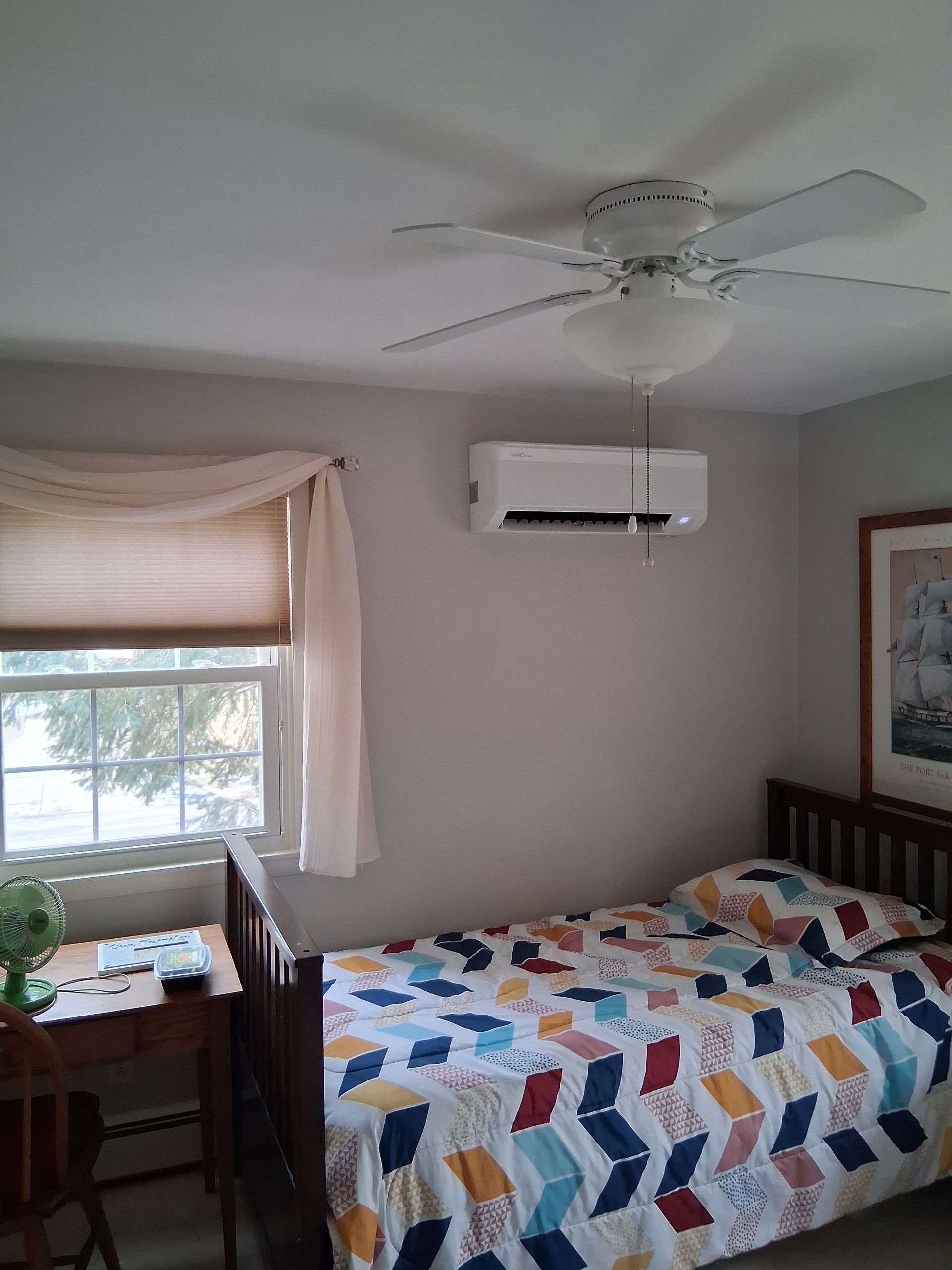 A bedroom with a bed featuring a colorful chevron pattern quilt, a window with beige curtains, and a ceiling fan.