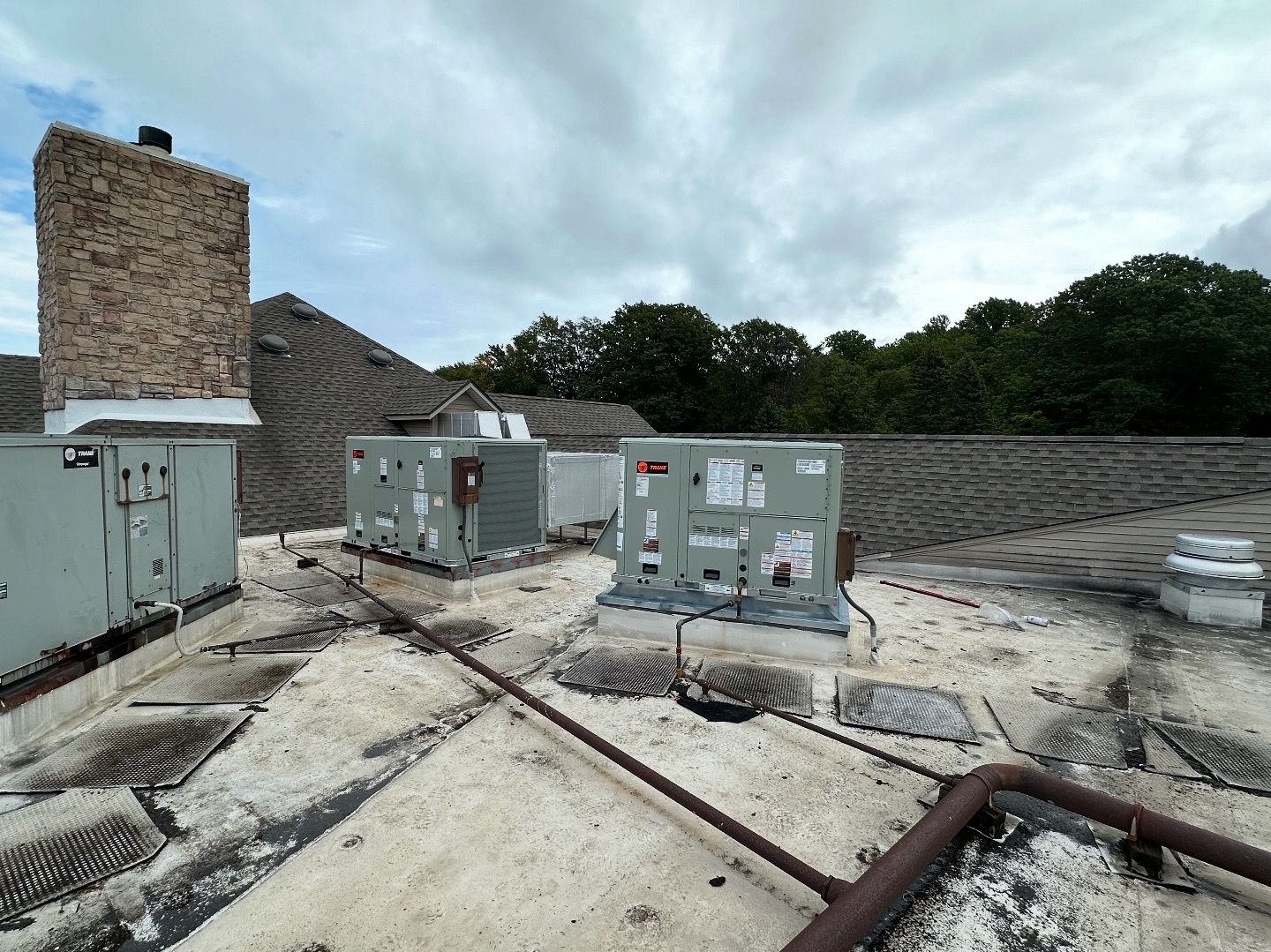 A flat commercial roof with multiple HVAC units, pipes, and a stone chimney under a cloudy sky.
