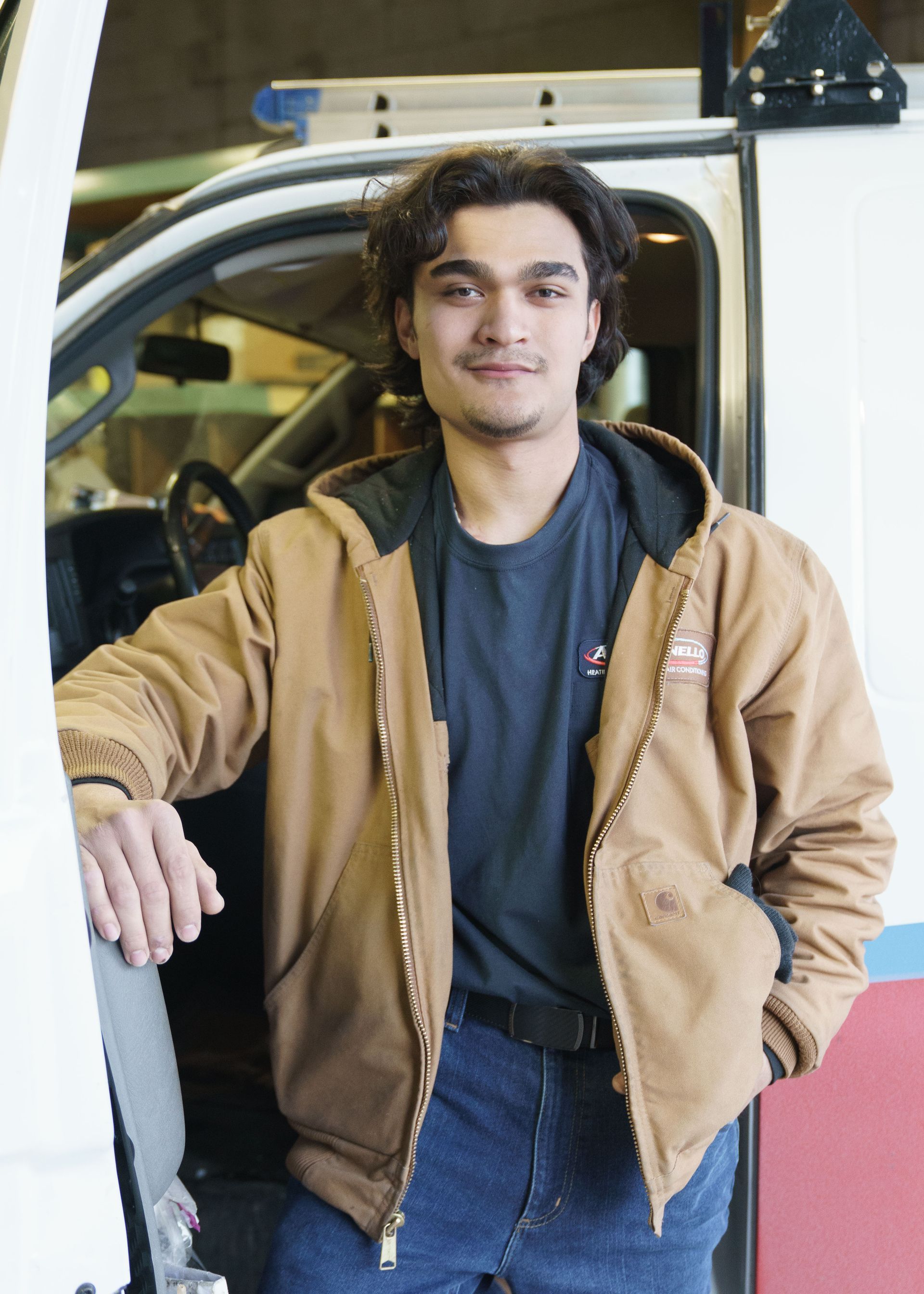 A smiling person wearing a tan jacket and dark shirt stands leaning against the open door of a white service vehicle.