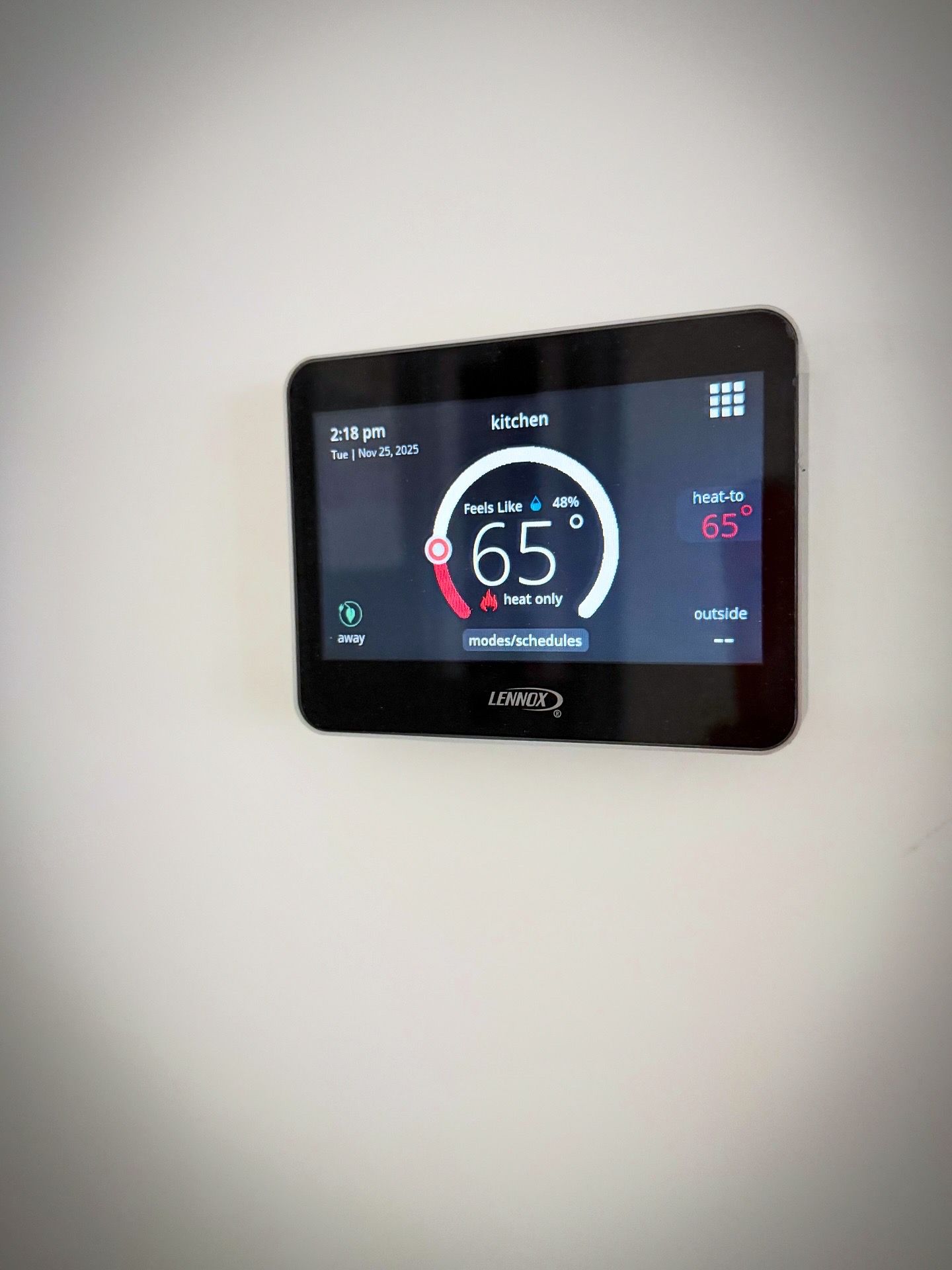 A black smart thermostat display mounted on a white wall shows a temperature of 65 degrees with digital interface controls.