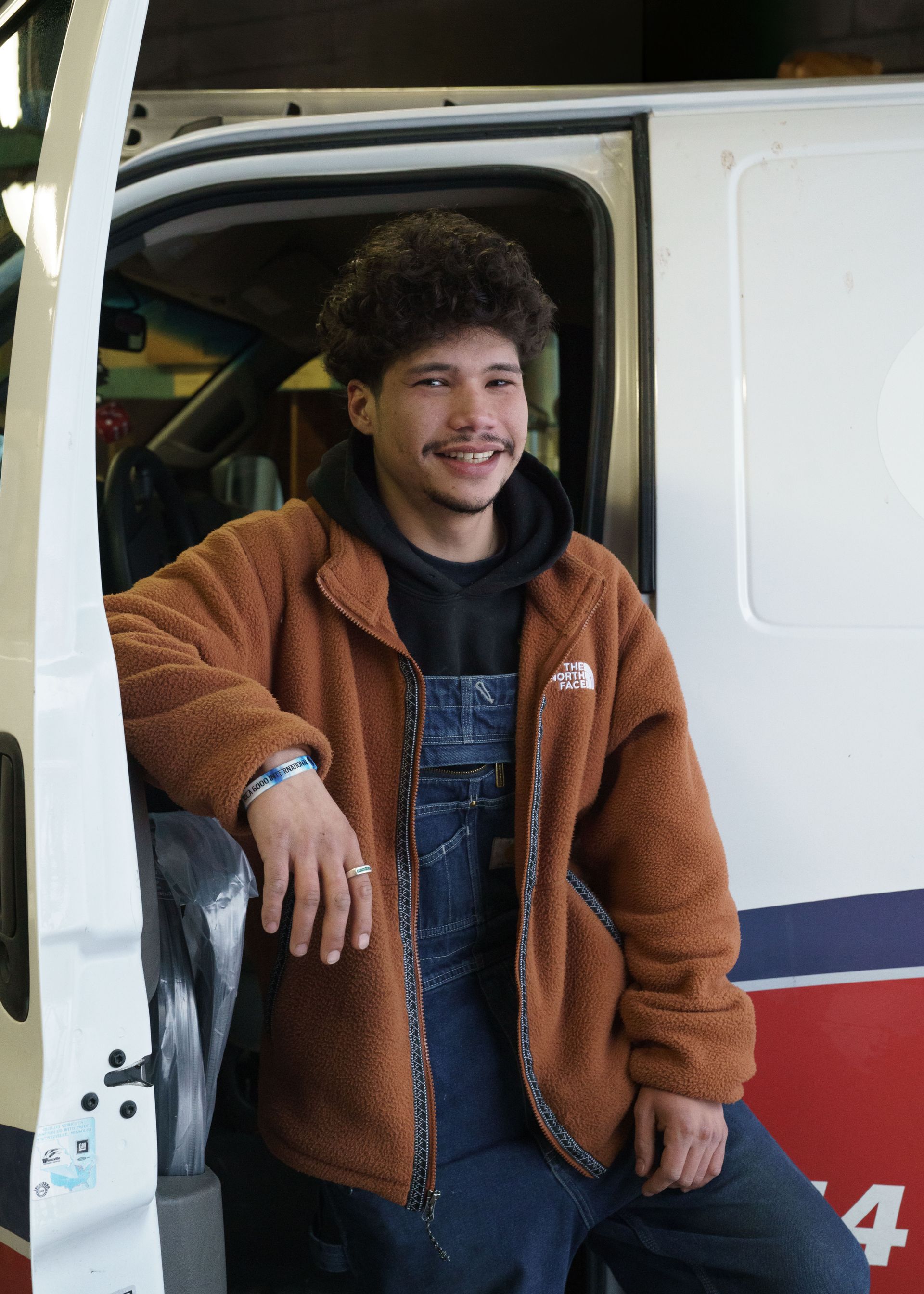 A smiling person in a brown fleece jacket and denim overalls leaning against the open door of a white van.