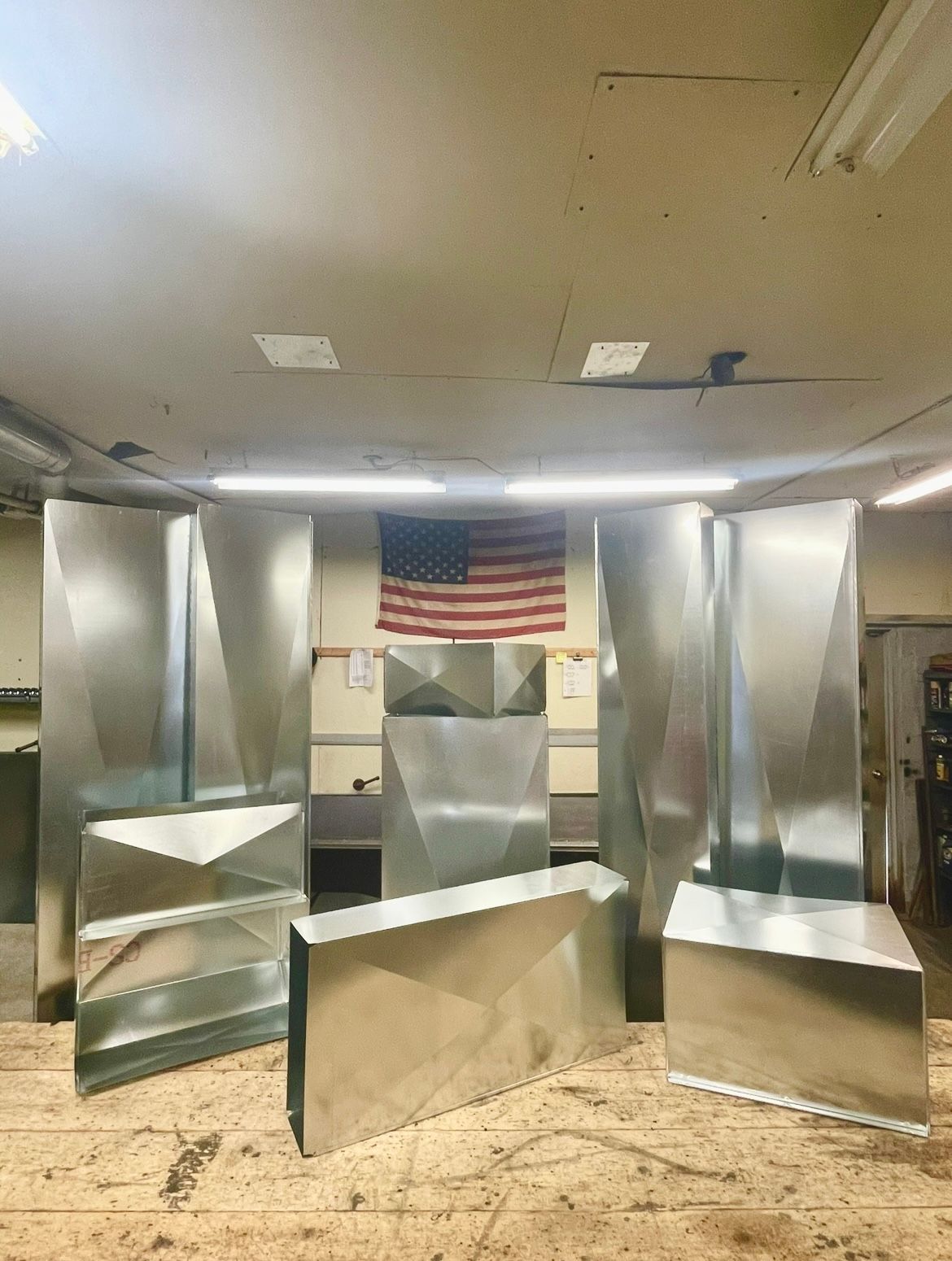 Various galvanized sheet metal HVAC duct components arranged on a workshop floor in front of an American flag.