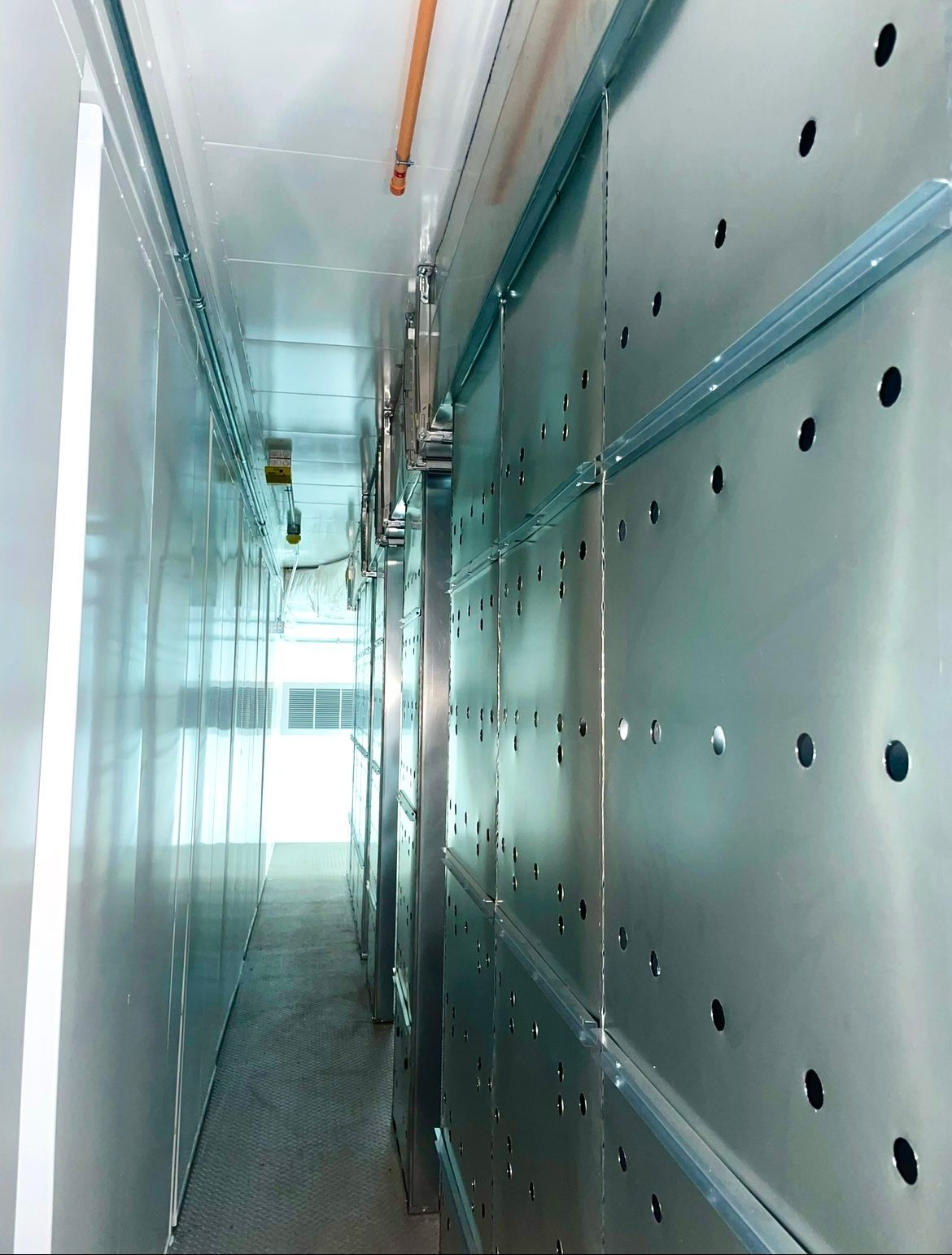 A narrow, industrial corridor with reflective, perforated metal walls and ceiling panels, brightly lit from the far end.