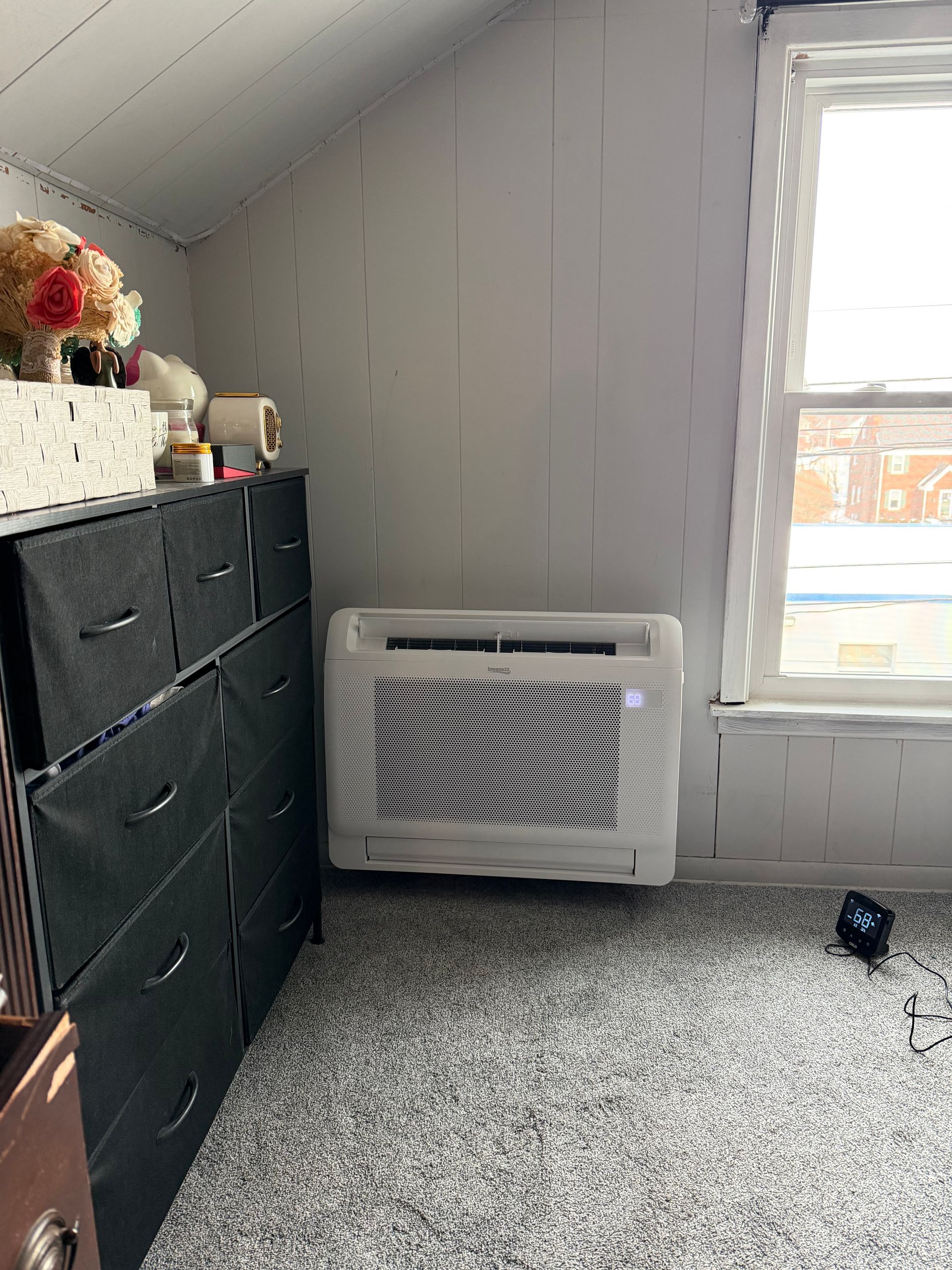 A black dresser sits beside a white wall-mounted air conditioning unit in a room with a sloped ceiling and window.