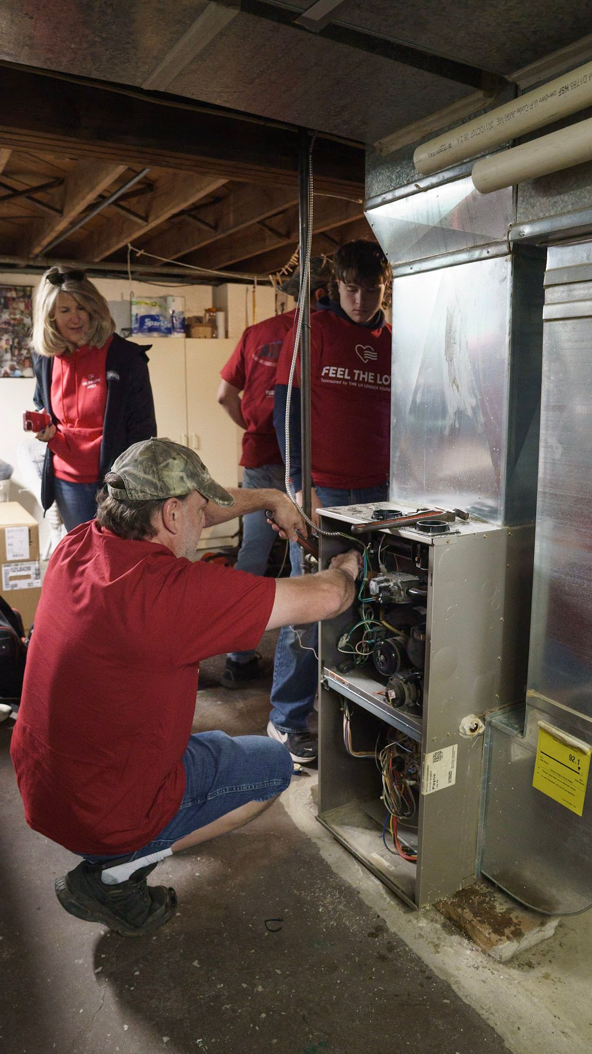A person in a red shirt kneels to repair an open furnace in a basement while two others observe.