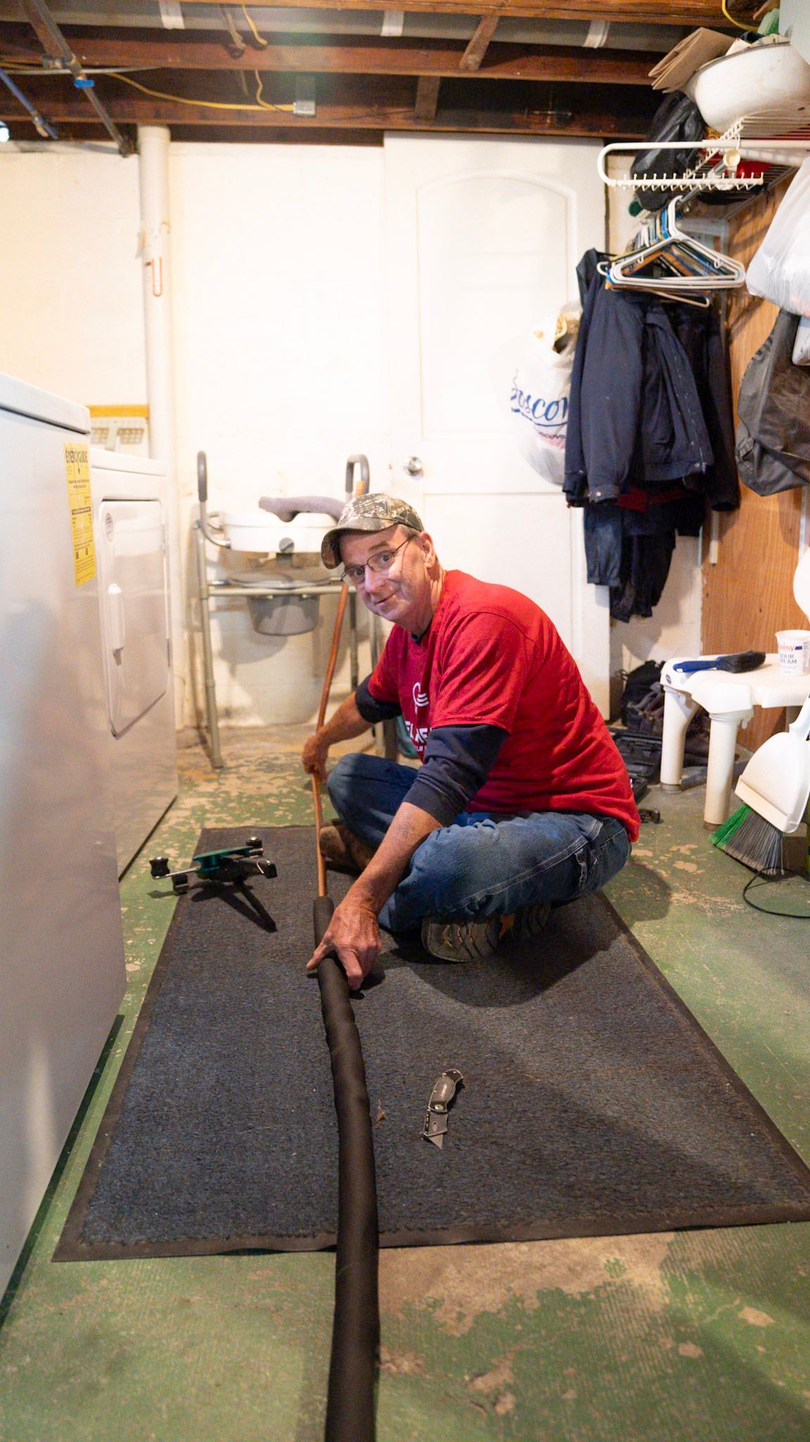 A person in a red shirt kneels on a basement floor, working with a long, dark pipe on a mat near a laundry machine.