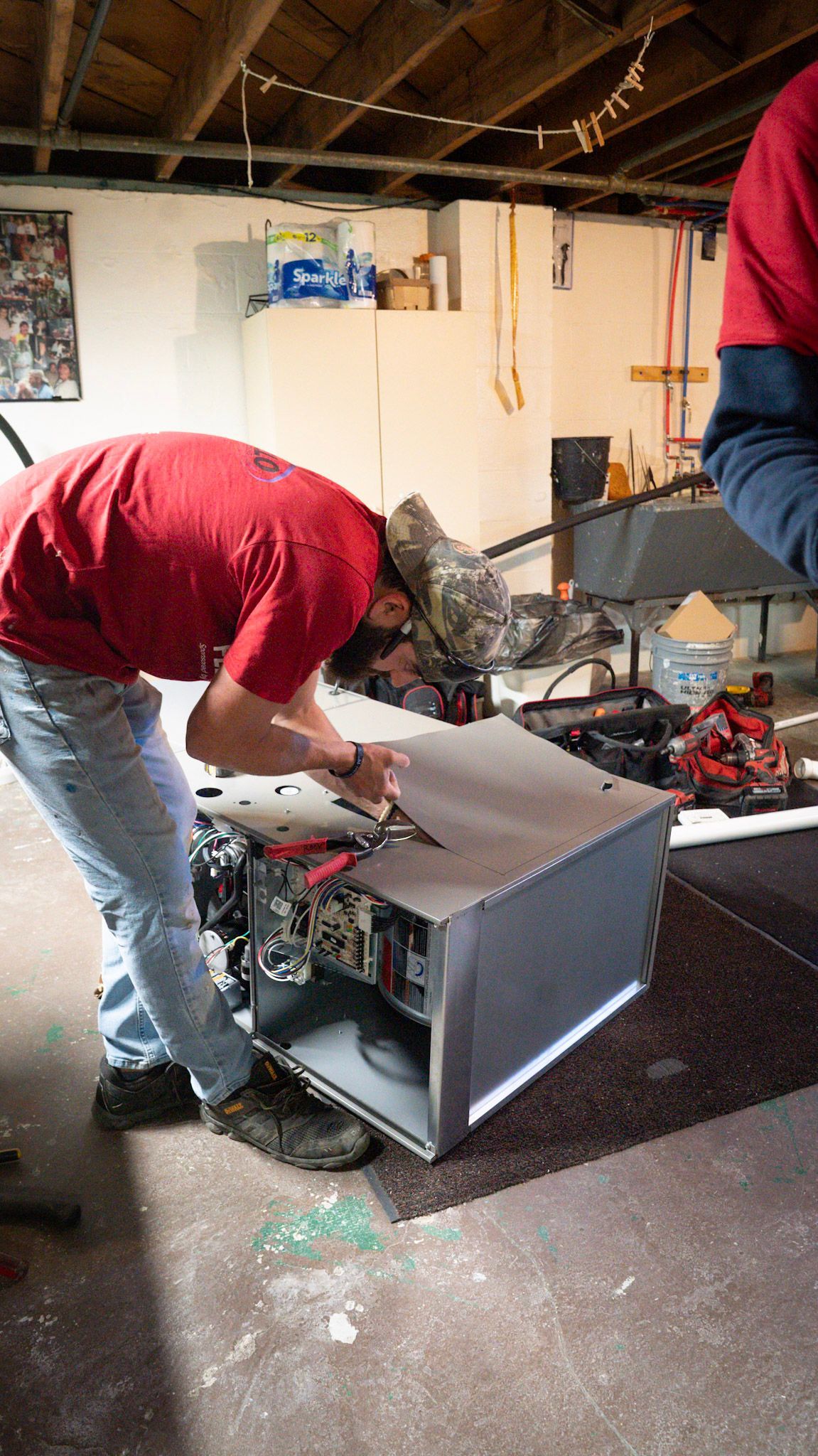 A person in a red shirt works on an open metal appliance in a basement setting.