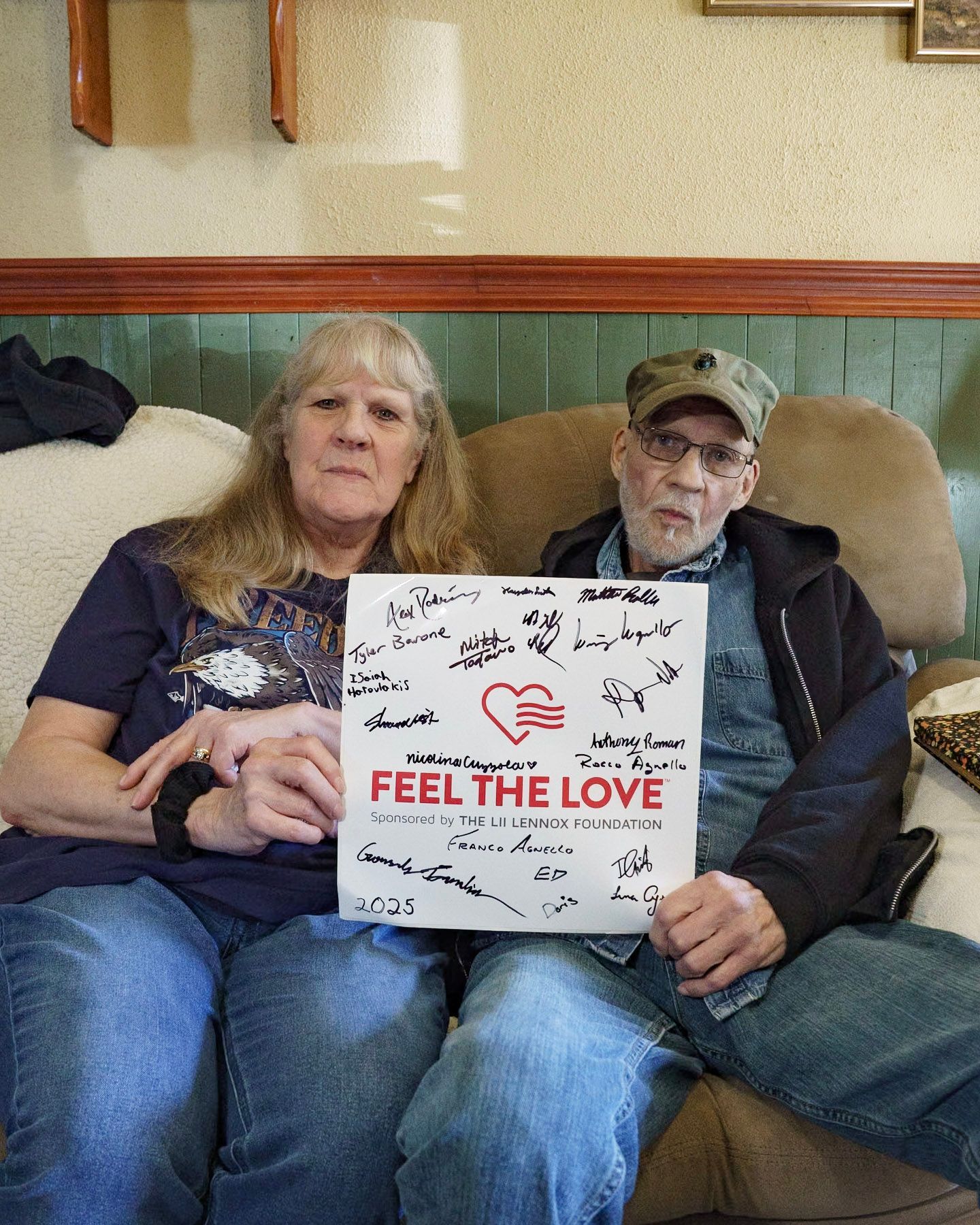 Two people sit on a sofa holding a sign that says 