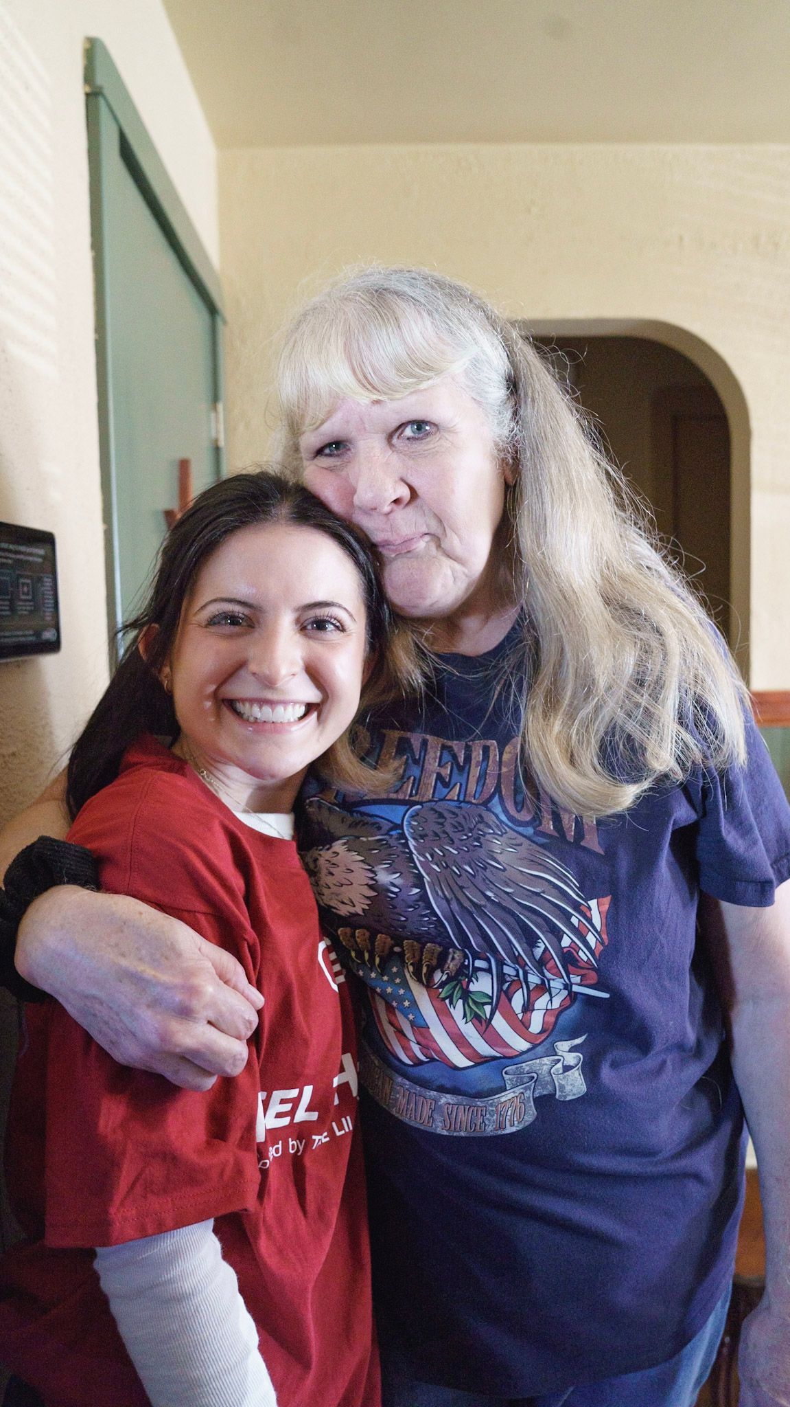 Two people stand together, smiling, with one arm around the other, in an indoor setting.