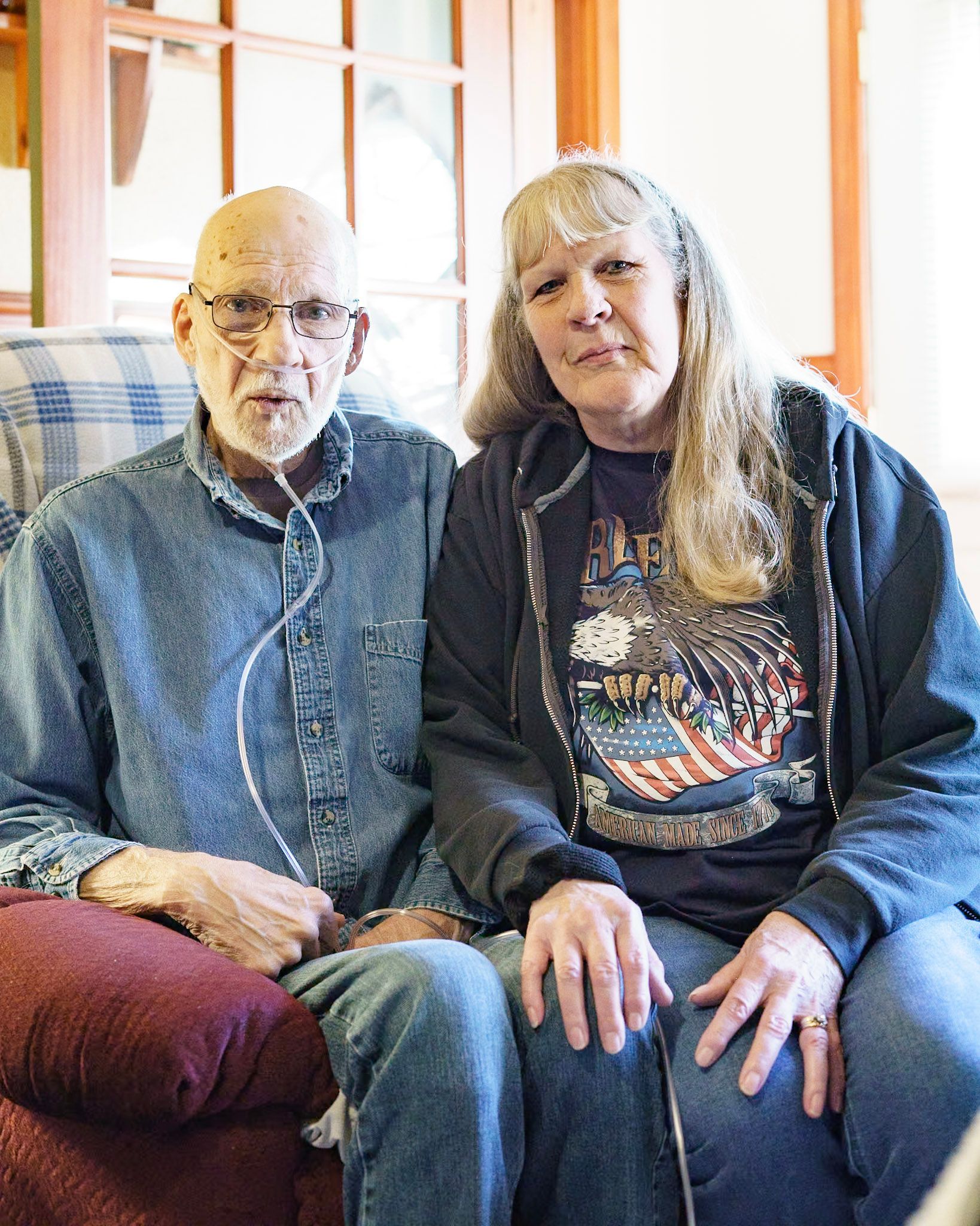 An older man wearing oxygen tubing and a denim shirt sits next to a woman in a dark graphic t-shirt and zip-up hoodie.