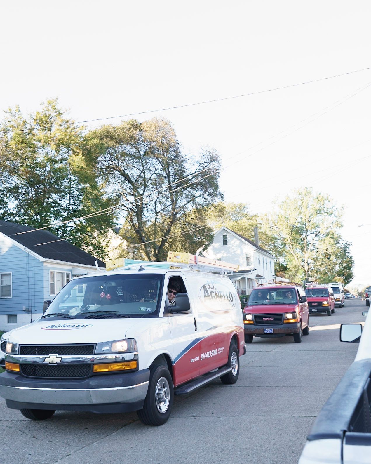 A white and red commercial van drives on a suburban street, followed by a line of other vehicles in daylight.