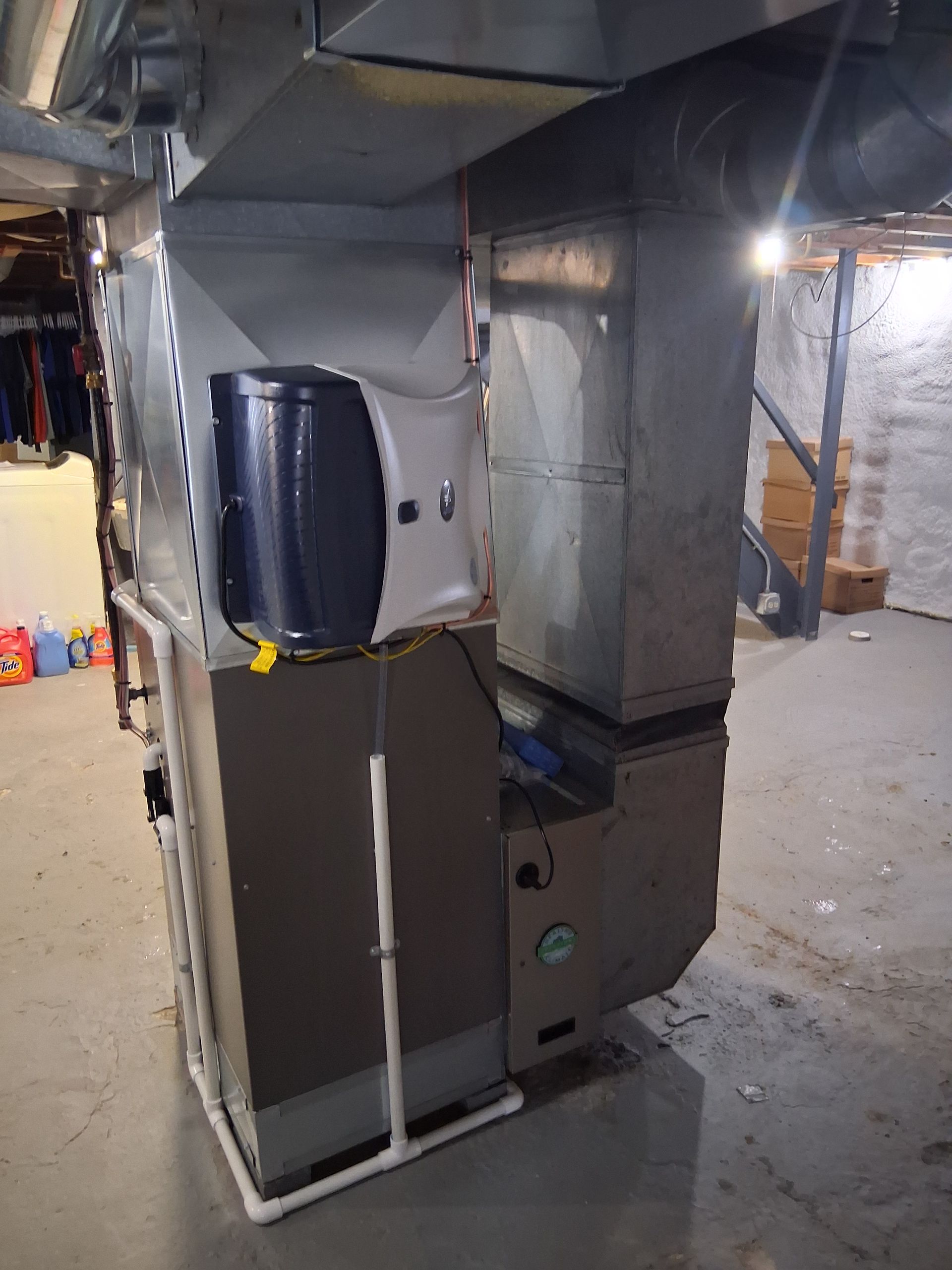 A basement HVAC furnace unit with an attached white and blue humidifier, connected to metal ductwork and a white PVC drain.