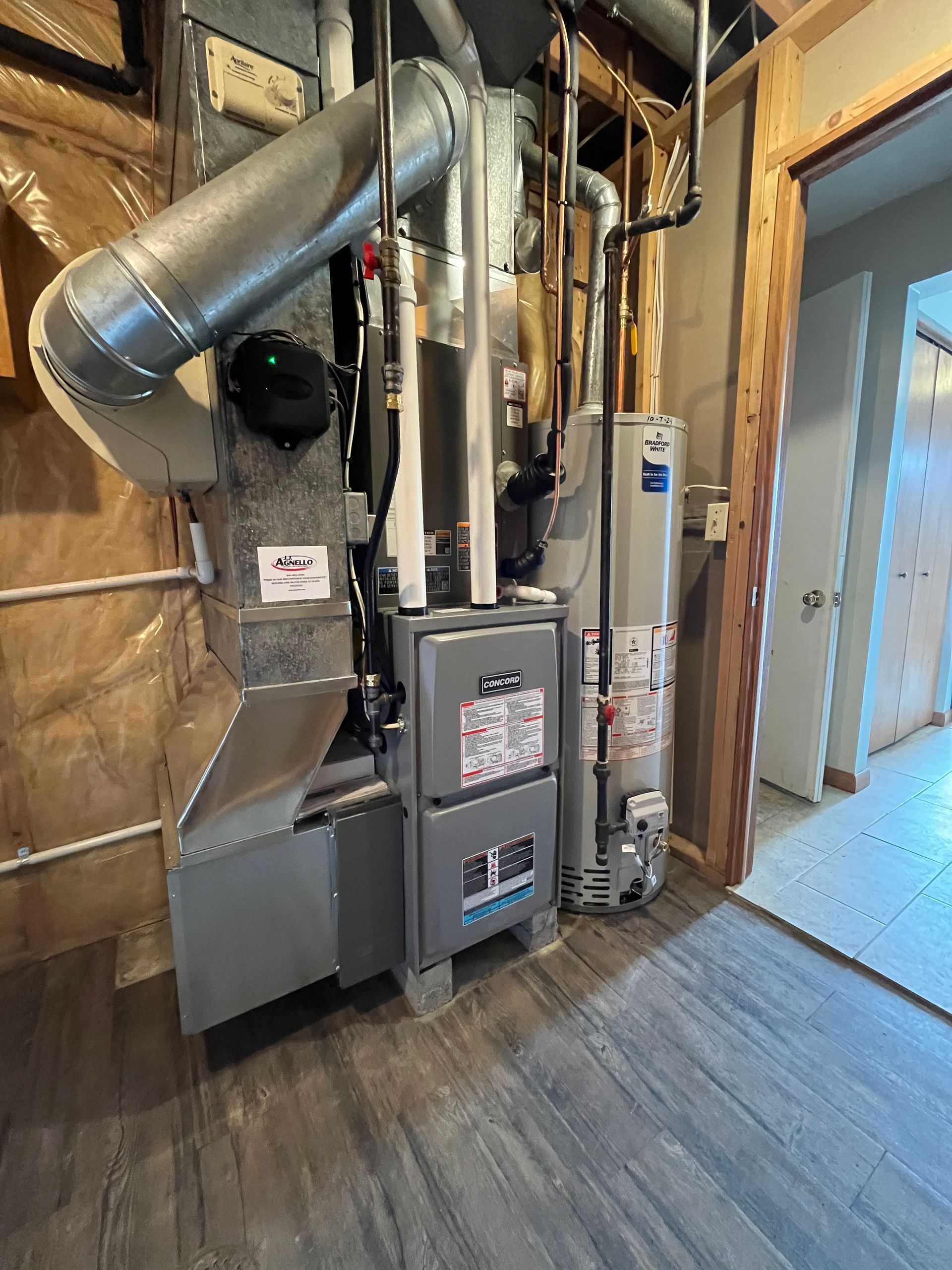 A furnace and water heater unit stand in an unfinished room with wood framing and grey flooring.