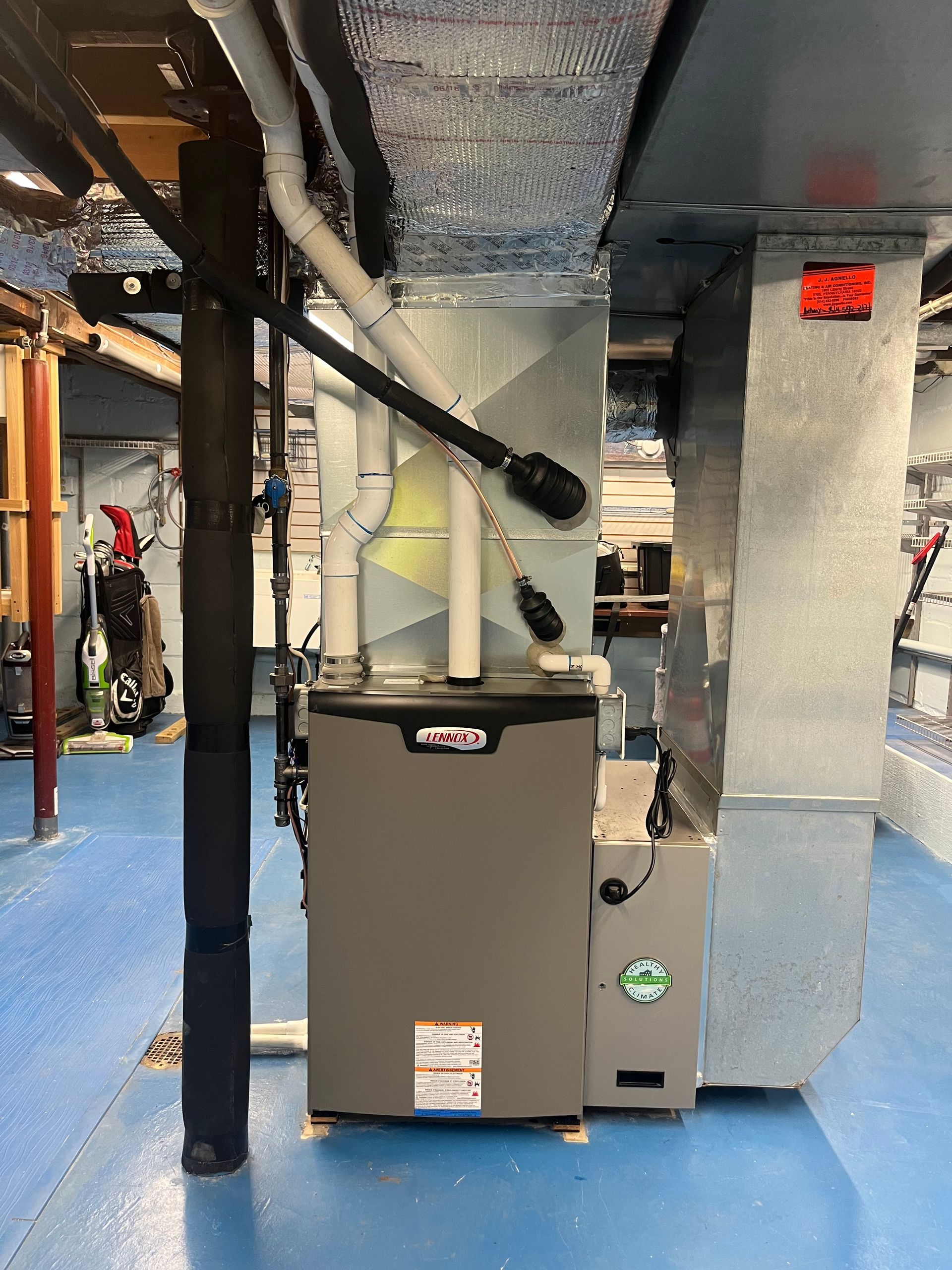 A grey furnace with white PVC pipes stands on a blue floor in a basement, connected to a metal ductwork system.