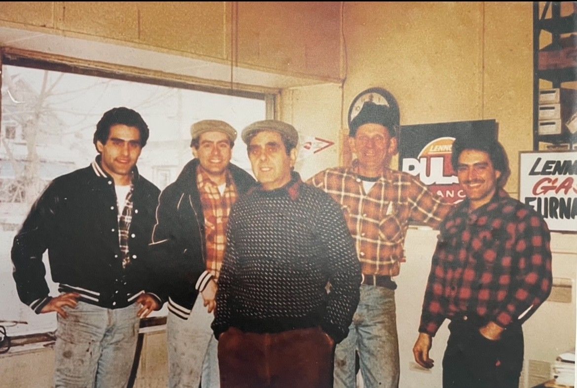 Five men pose indoors by a window. One smokes. A sign reads 