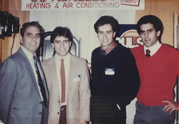 Five men standing indoors near a window, two with caps, one with a mustache, all wearing casual clothes.
