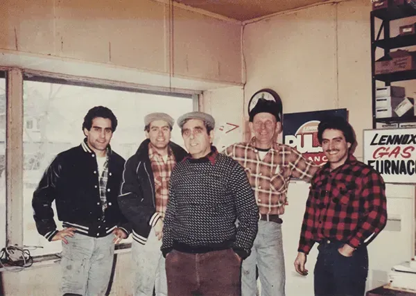 Five men standing indoors near a window, two with caps, one with a mustache, all wearing casual clothes.