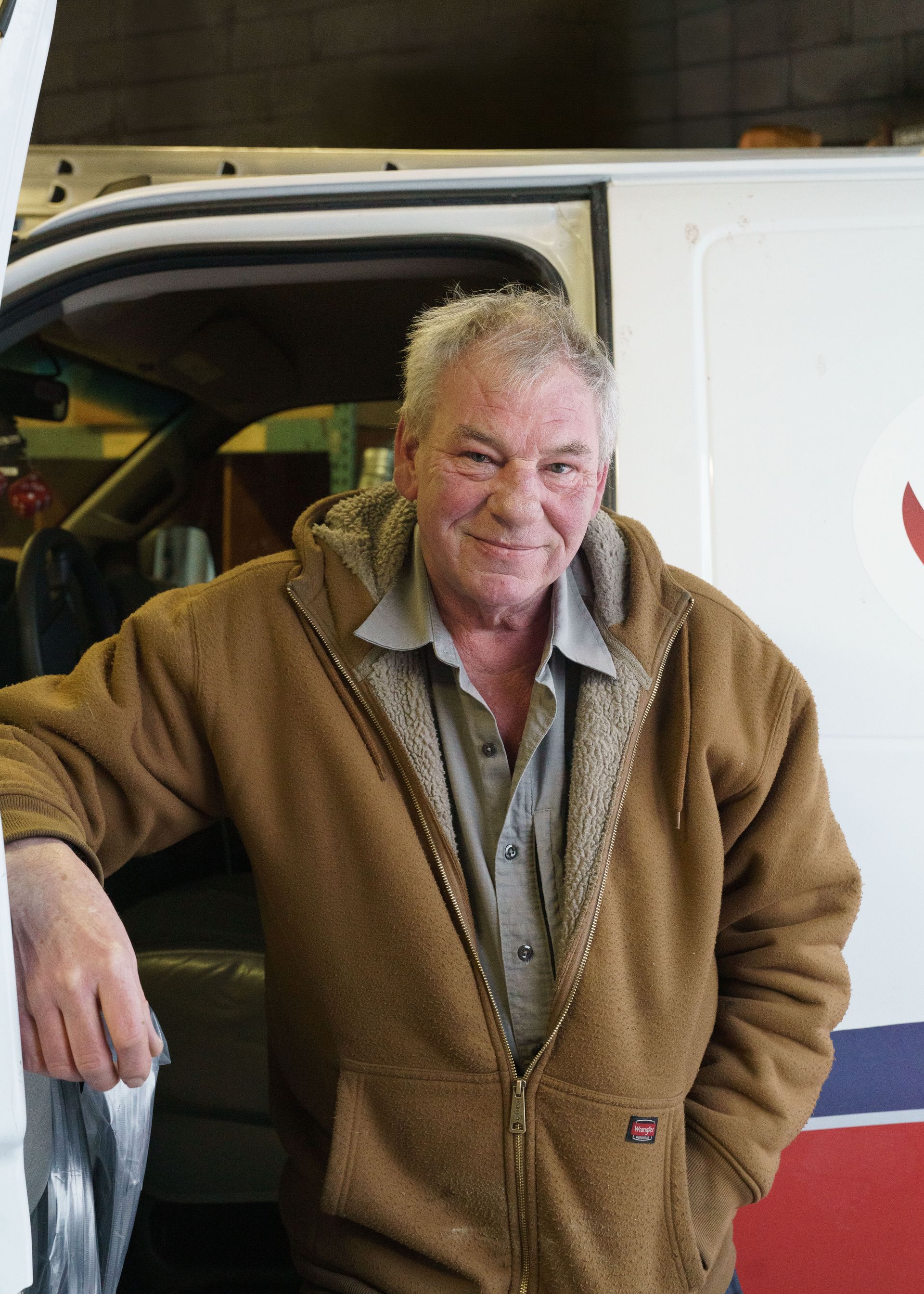 A person in a brown fleece hoodie smiling while leaning against the open door of a white van in a garage.