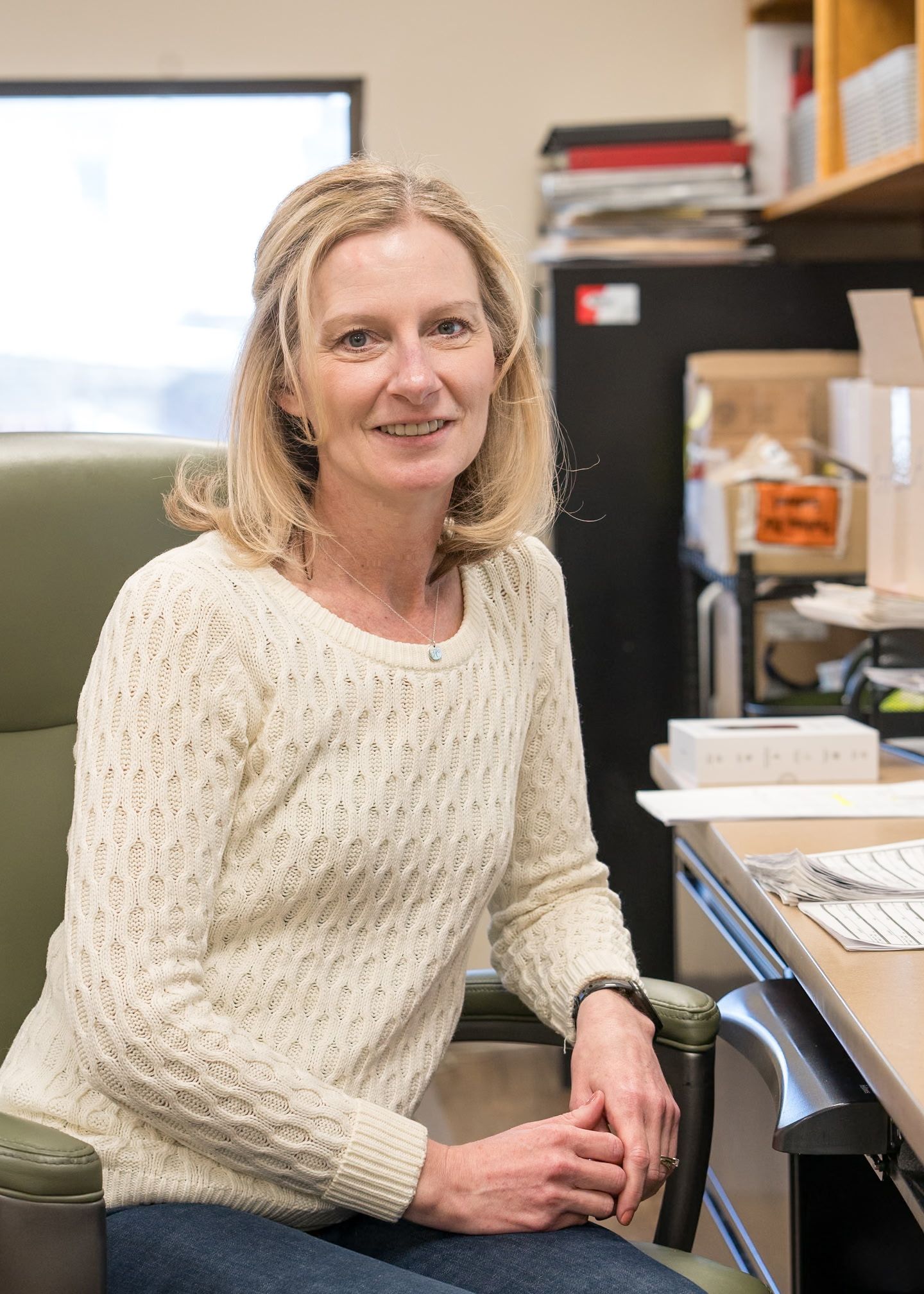 A person with shoulder-length blonde hair sits at a desk in an office, wearing a cream-colored knit sweater.