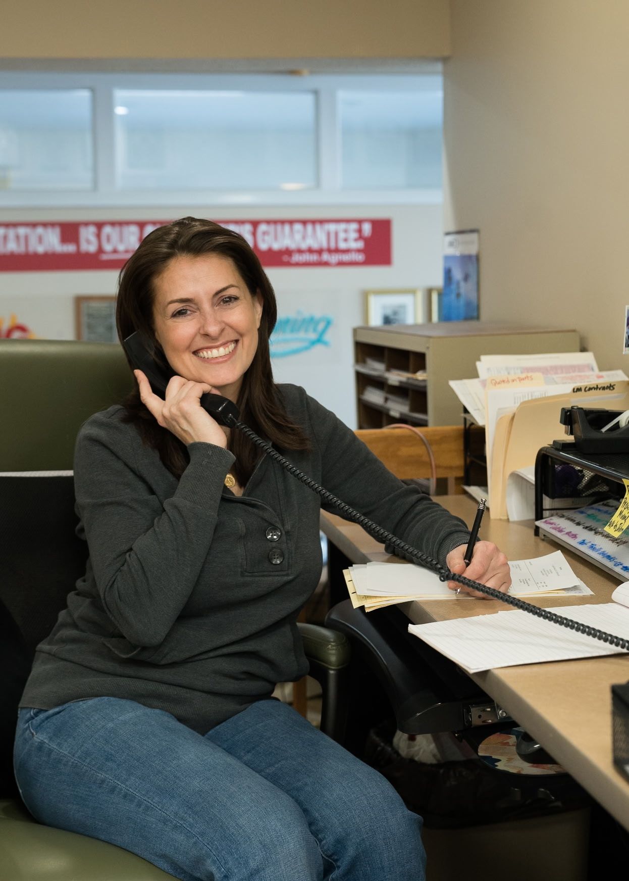 A smiling office worker holding a telephone receiver to her ear while holding a pen over a desk with paperwork.