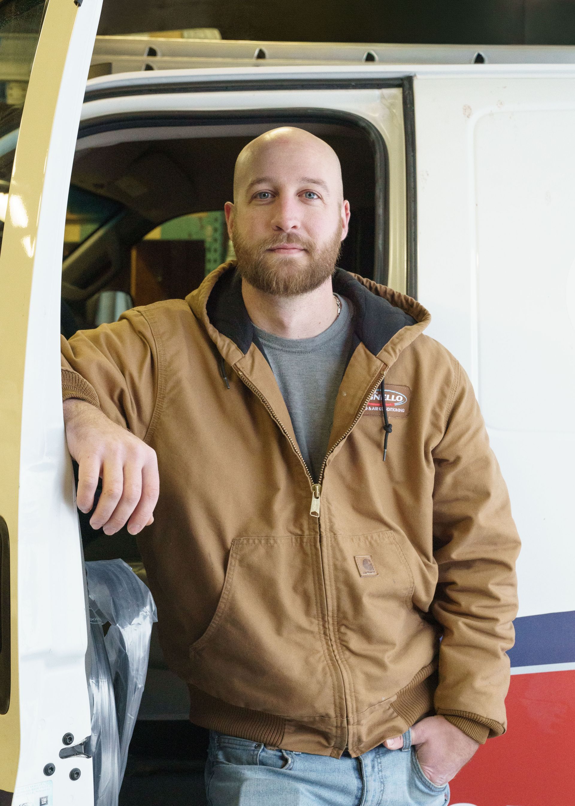 A person with a beard, wearing a brown hooded work jacket and grey shirt, stands next to an open white van door.