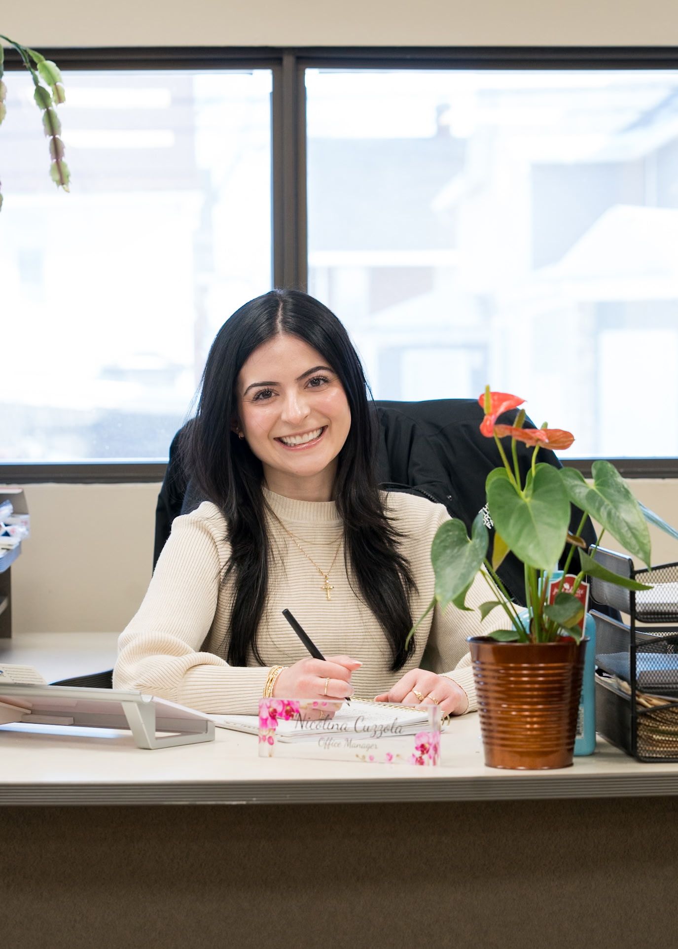 A person smiling while sitting at a desk with a potted anthurium plant, pen in hand, in a well-lit office setting.