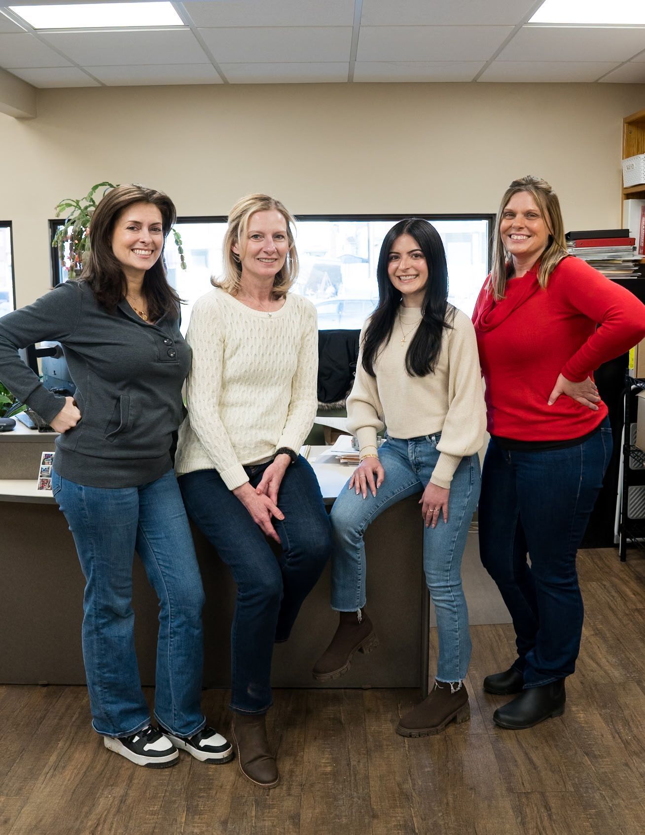 Four people pose smiling in an office, wearing casual clothing, standing and sitting near a desk.