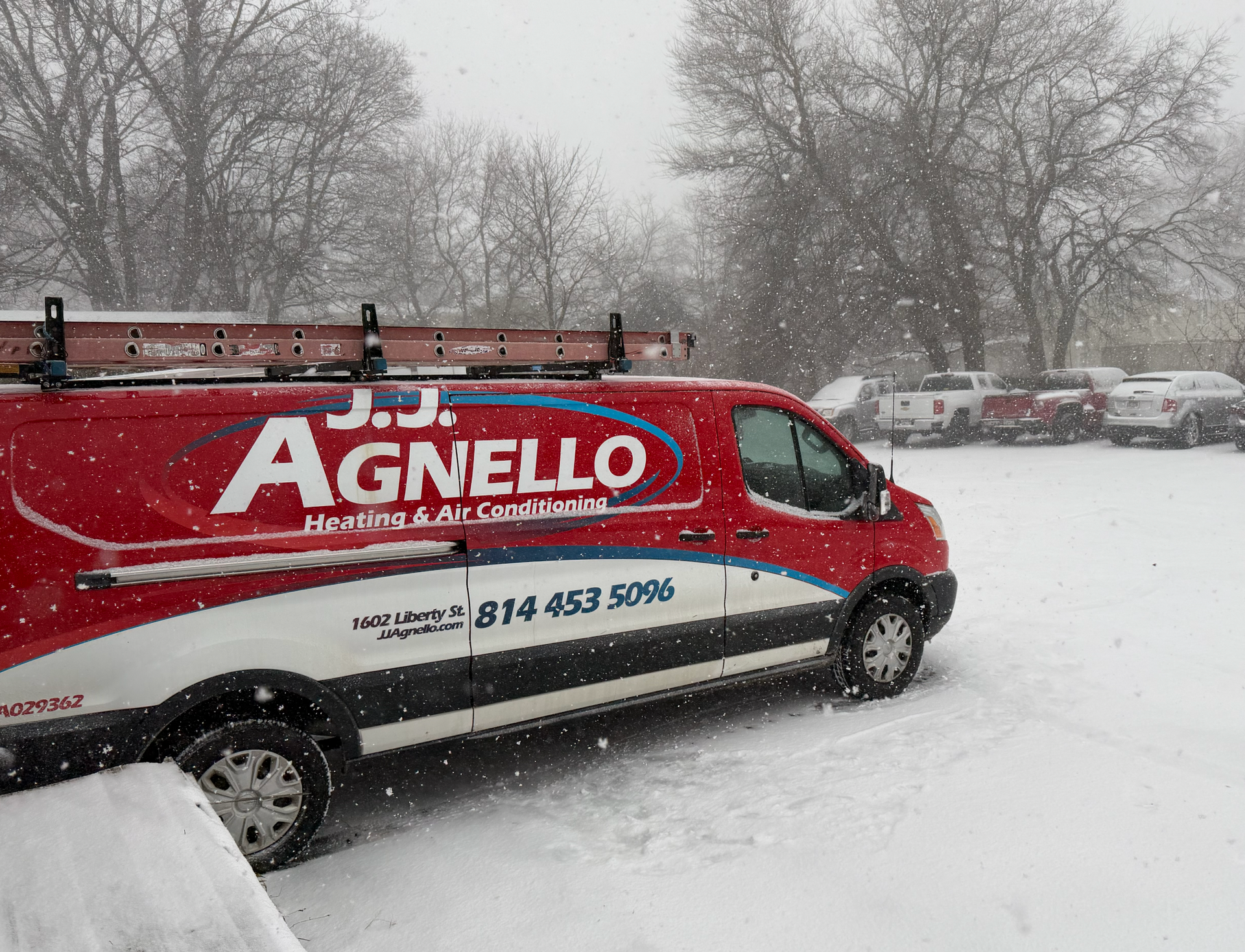 Red and white J.J. Agnello service van with ladder in snowy parking lot. Trees and cars in the background.