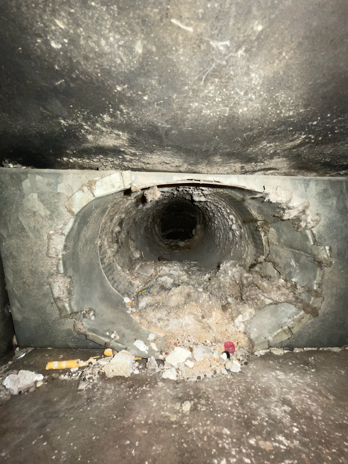 A dark, circular concrete pipe opening in a rough, grey wall, with debris and dust scattered on the ground below.
