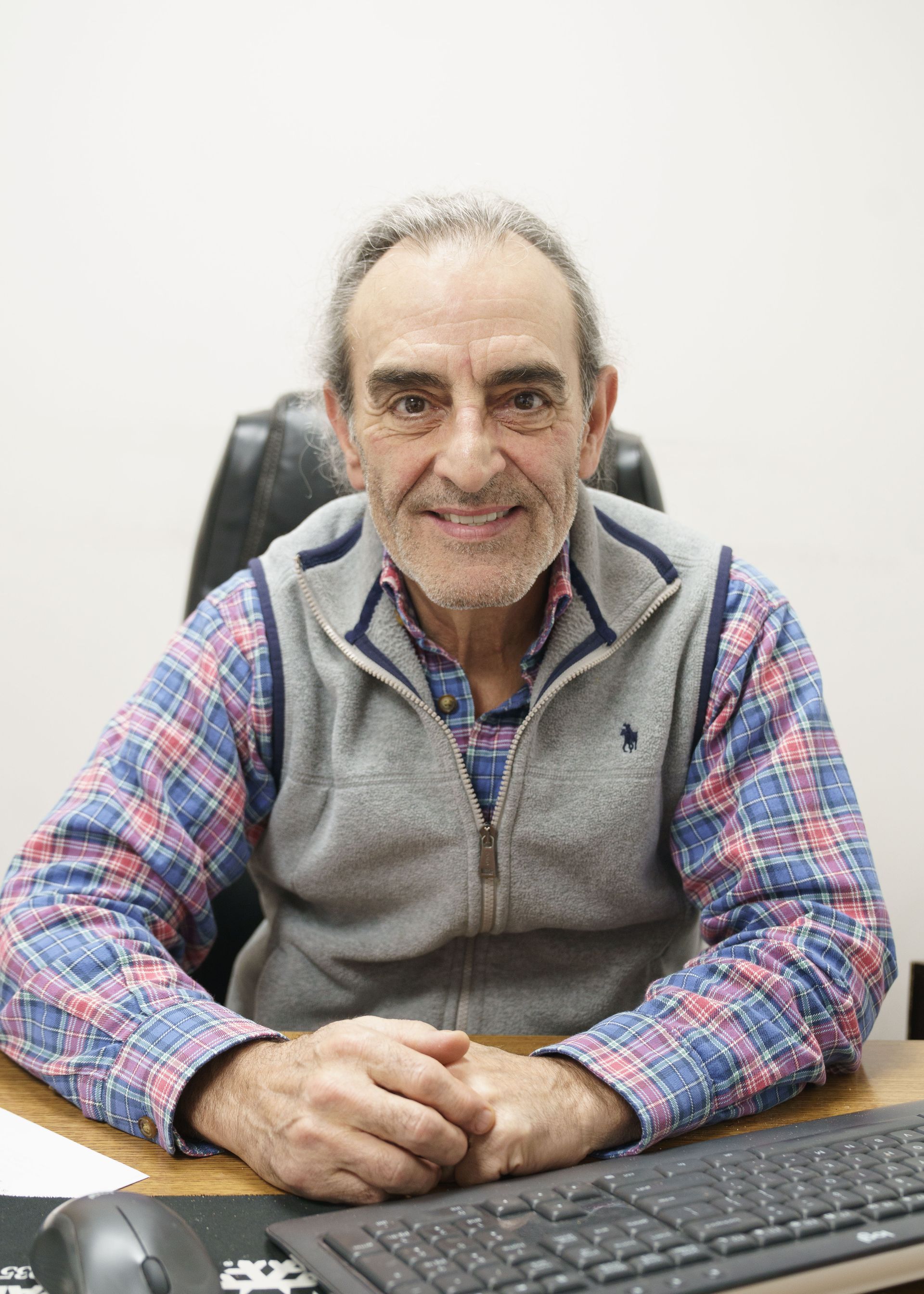 A smiling person sits at a desk with a keyboard, wearing a plaid shirt and a gray vest in an office setting.