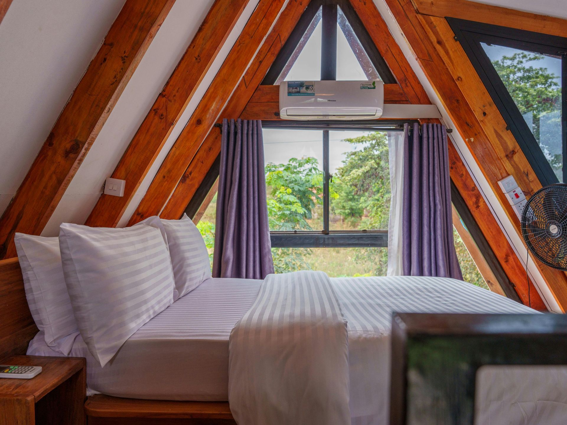 Bedroom with wooden A-frame ceiling, white bedding, window, curtains, air conditioner, and fan.