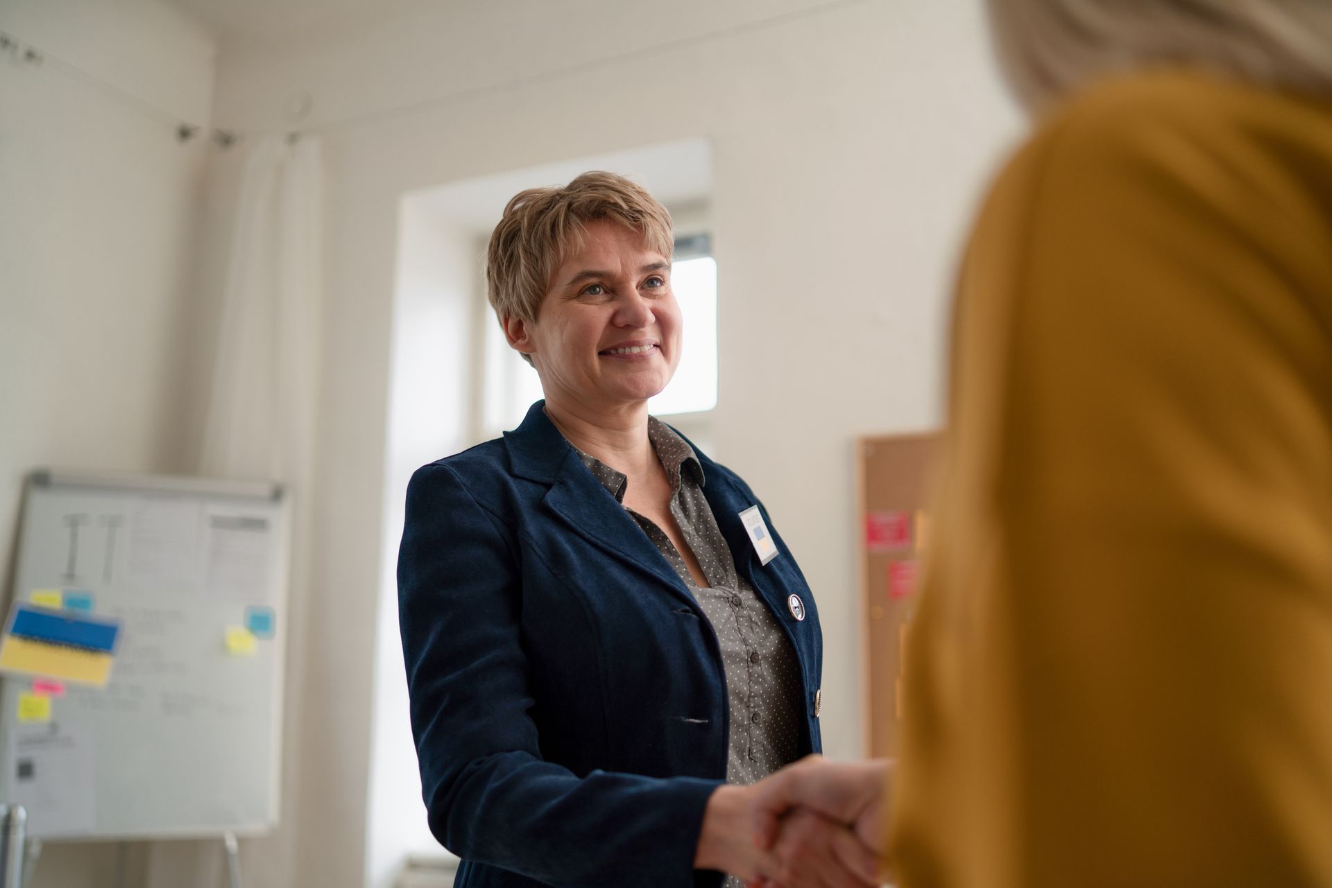 A woman in a blue jacket is shaking hands with another woman in a yellow shirt.
