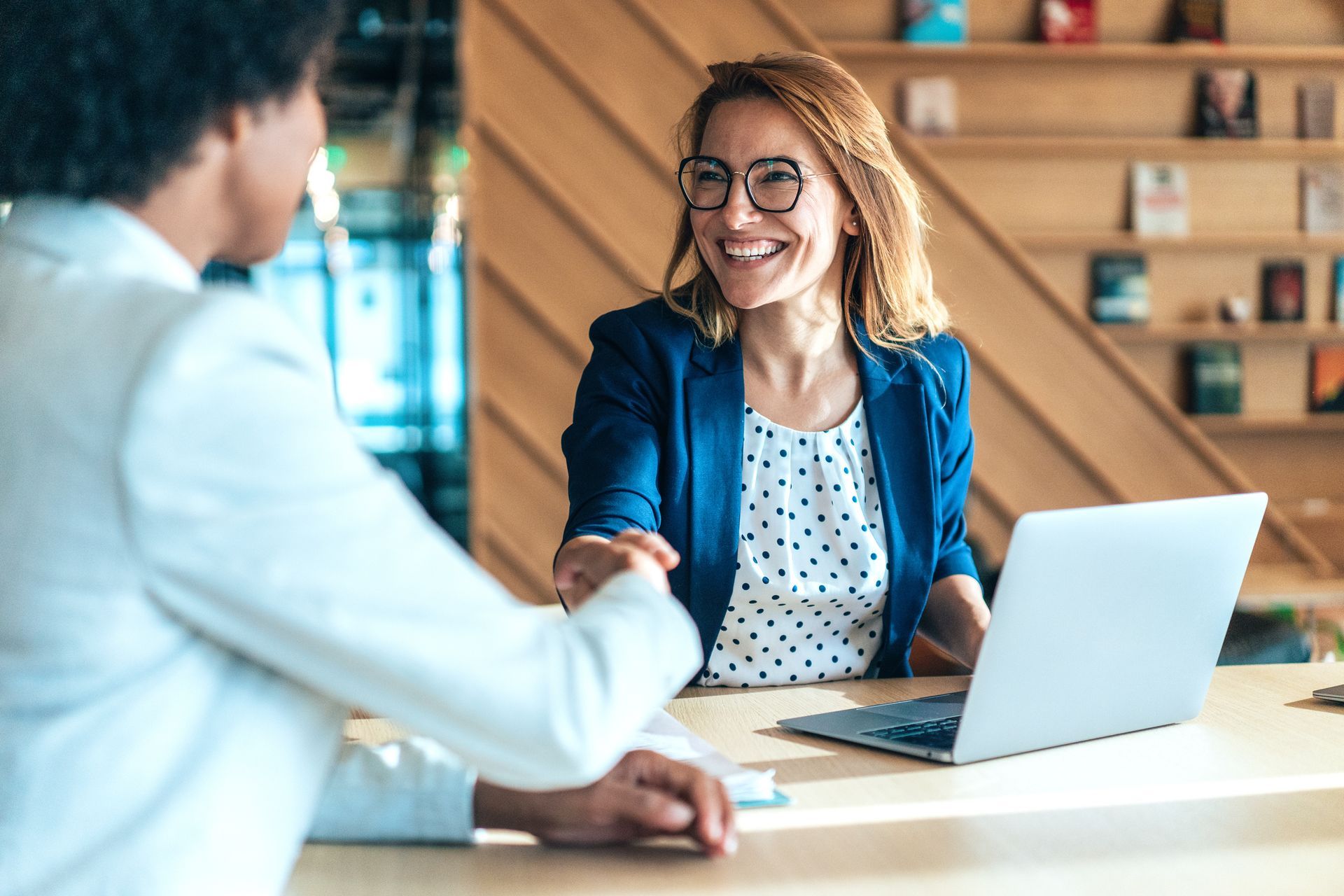 A woman is shaking hands with a man while sitting at a table with a laptop.