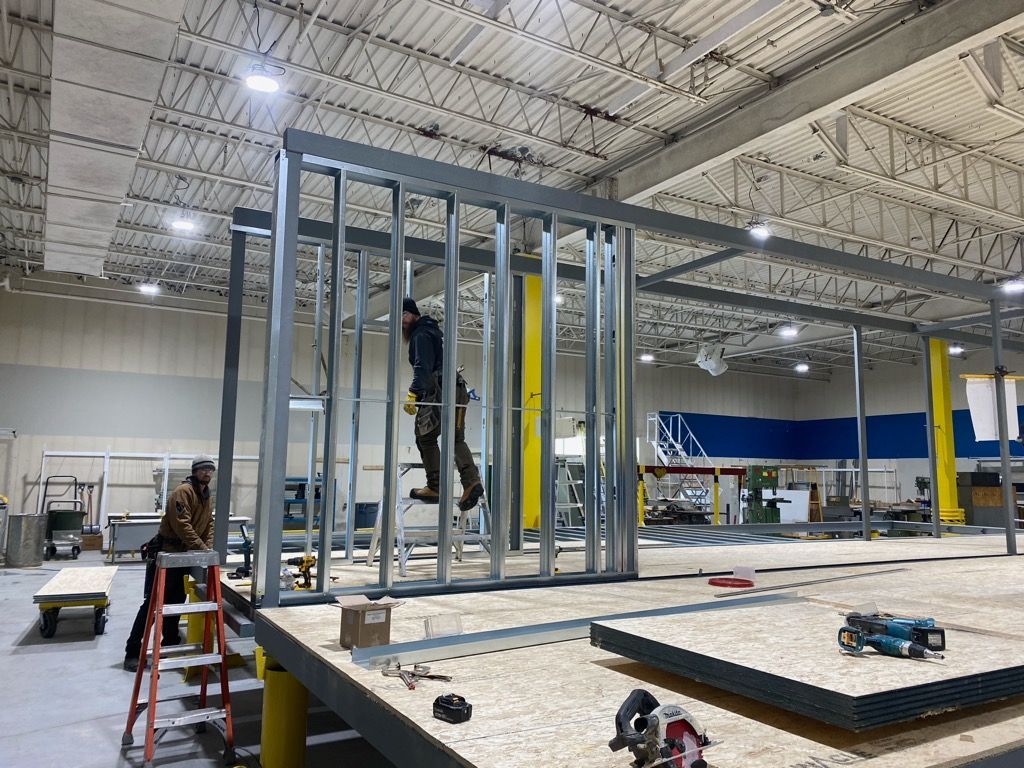 Two workers assemble a metal stud wall frame inside a large, high-ceilinged workshop with a wood-paneled floor.