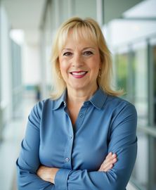 Woman with blonde hair, smiling, arms crossed, wearing blue shirt, standing in a modern building.